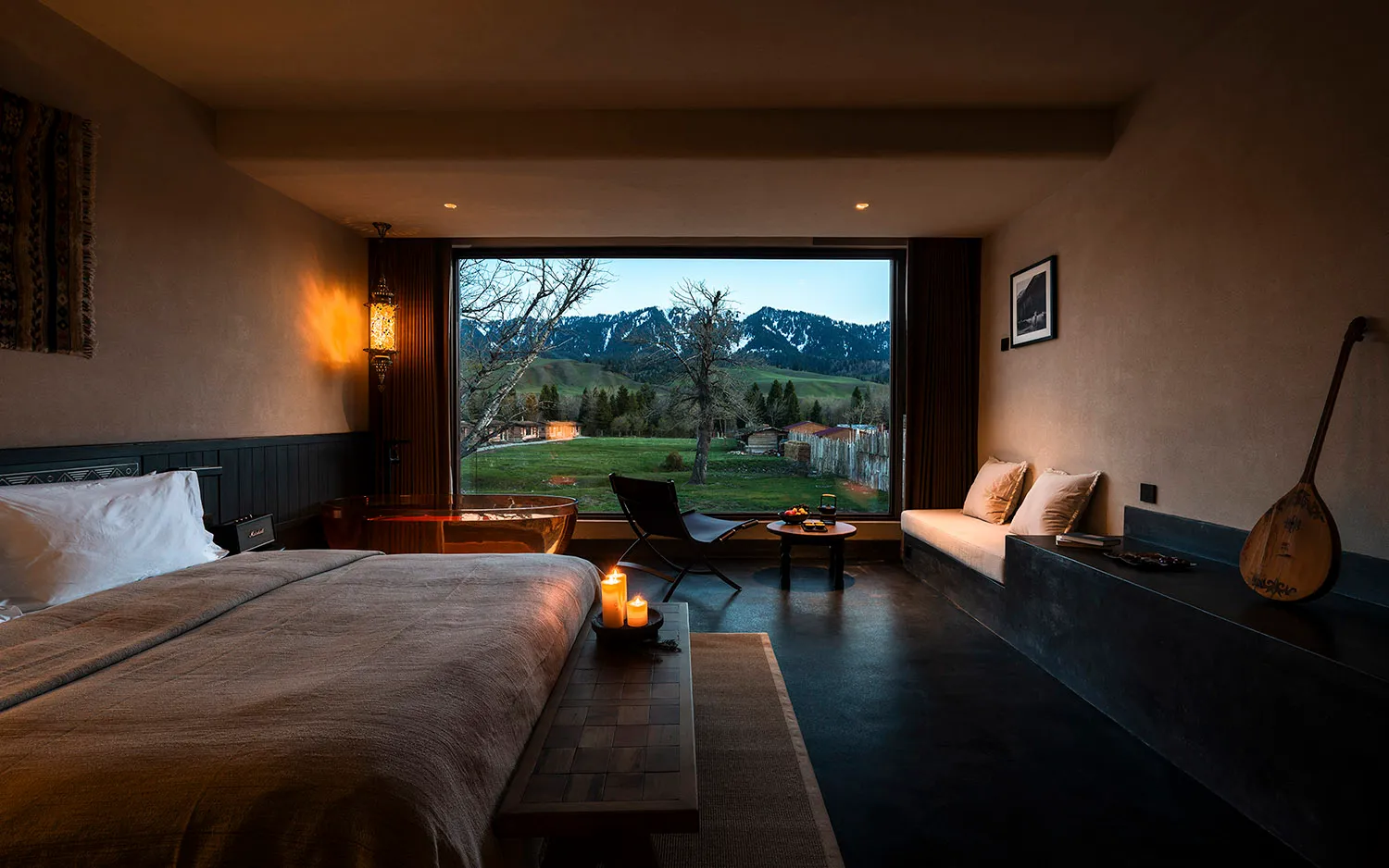 Modern bedroom interior featuring a large bed with taupe linen, a freestanding copper bathtub, built-in window seat, and dramatic mountain landscape visible through floor-to-ceiling windows.