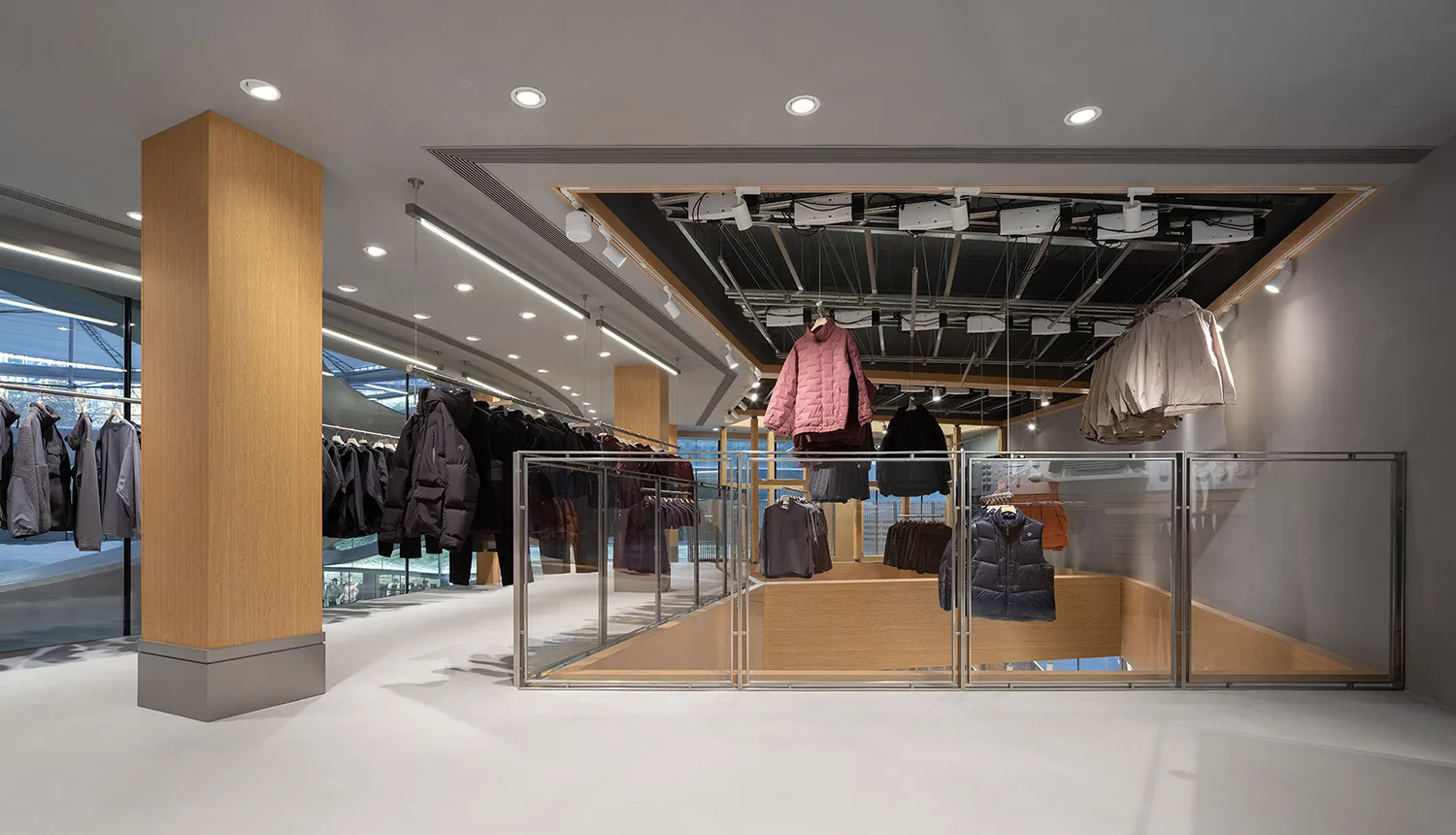 Modern minimalist clothing store interior with large oak columns, white floors, glass railings, and clothing hanging from an overhead industrial grid system under recessed lighting.