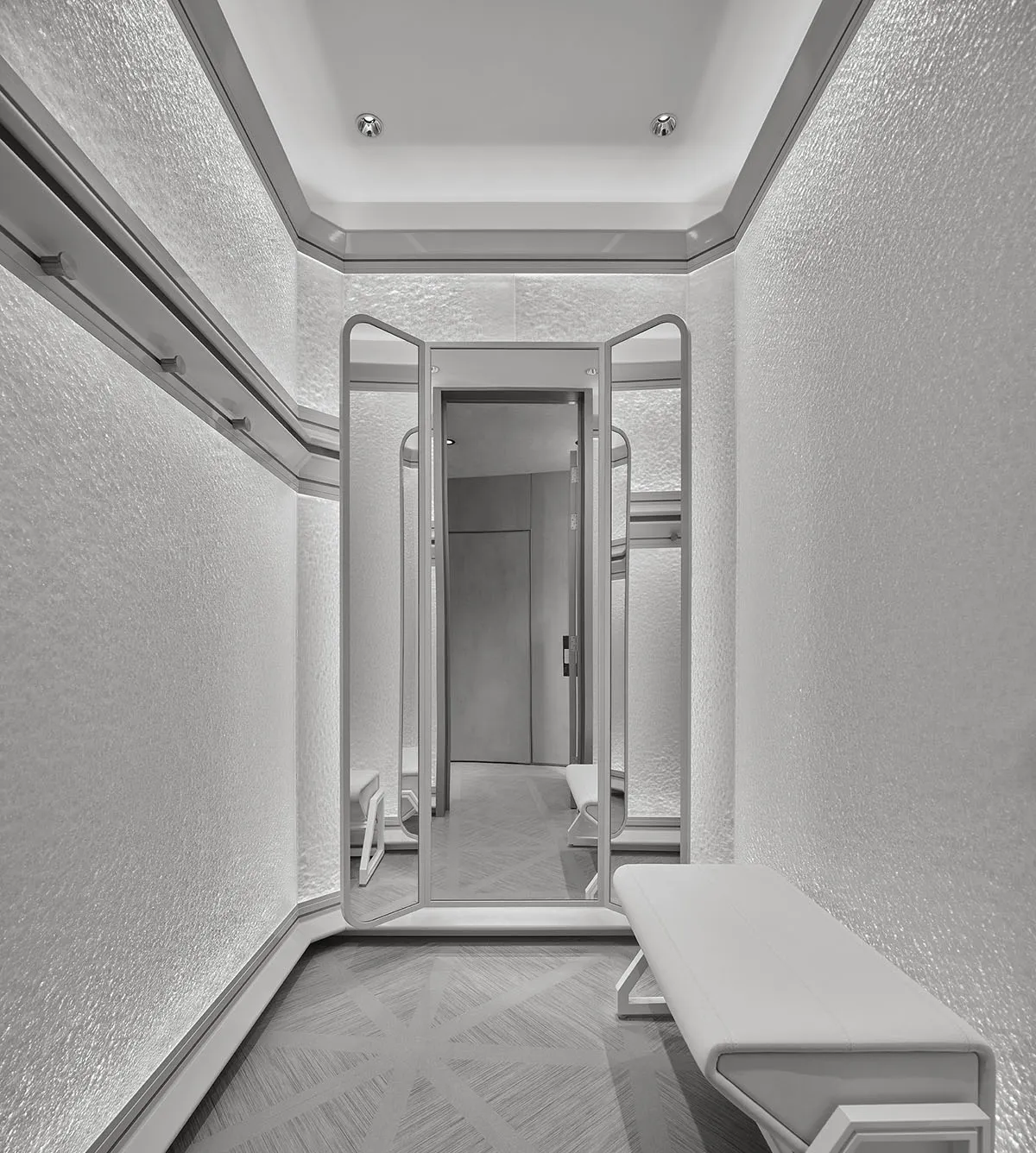 Modern minimalist dressing room with white textured plaster walls, geometric patterned carpet, white upholstered bench, and multi-panel mirrored doors reflecting the space.