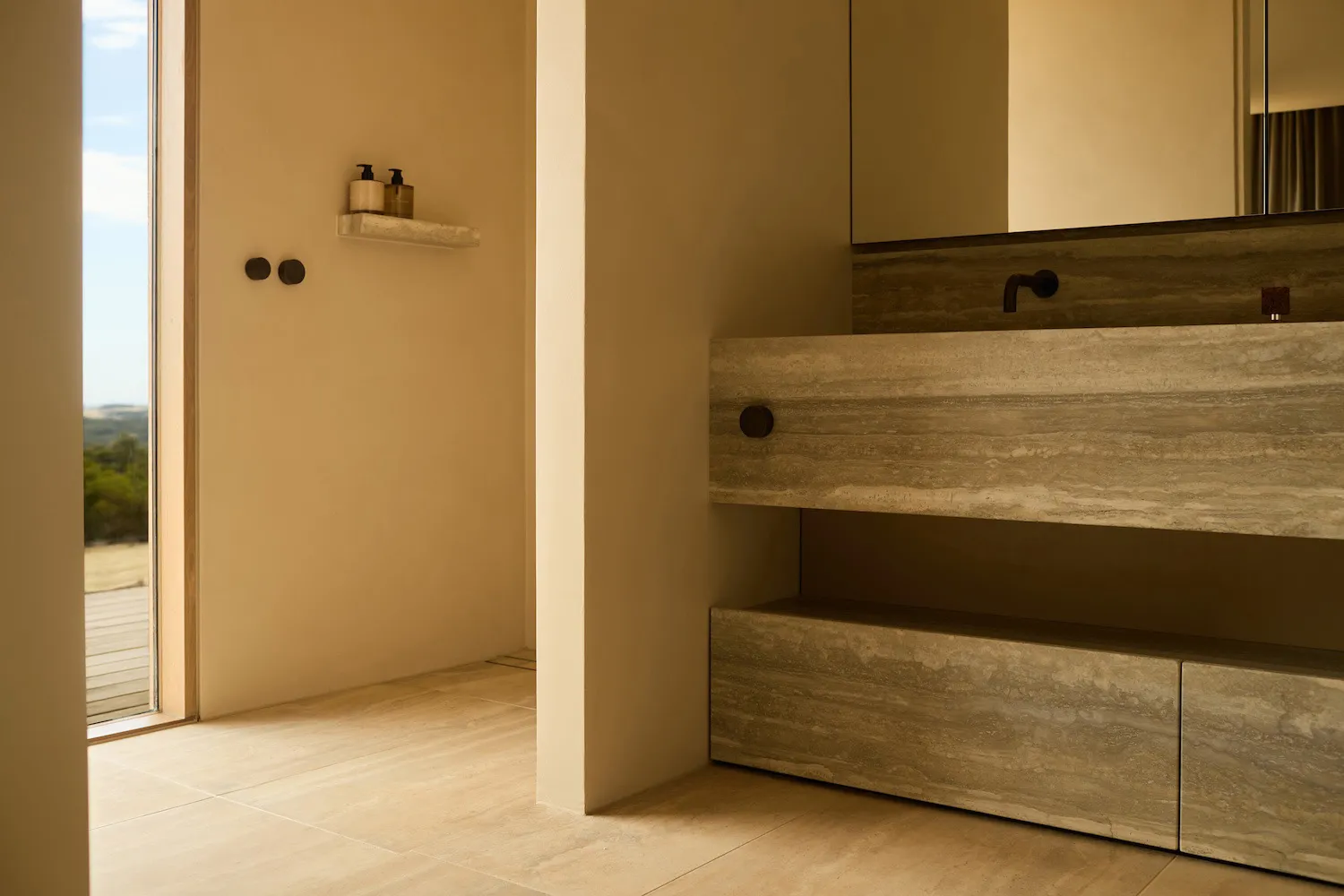 Modern minimalist bathroom featuring a floating travertine vanity with integrated mirror and black matte faucet, warm beige walls, and an open doorway revealing a scenic outdoor view.