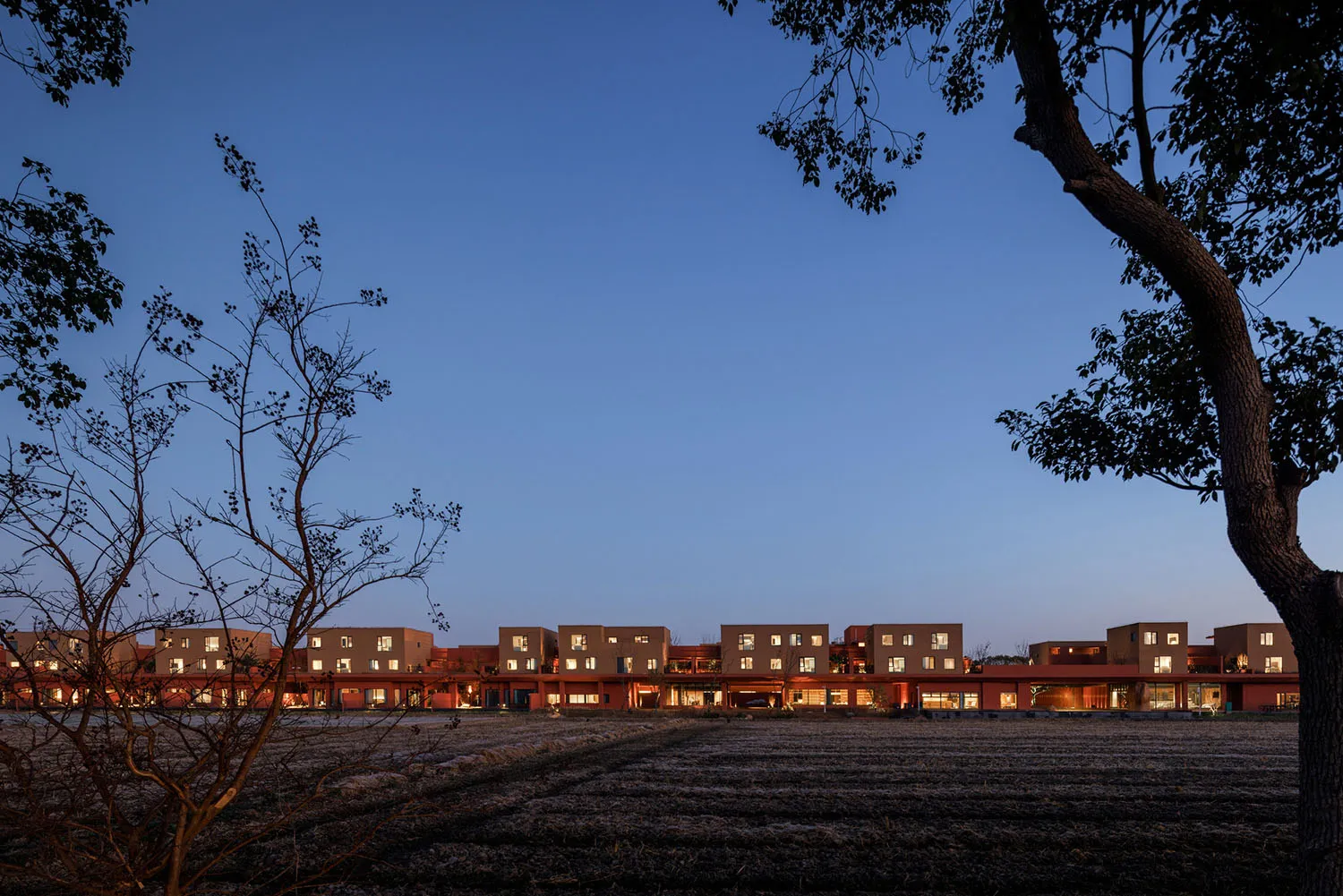 台灣鄉間收割後的田野前方，一排現代陶土色住宅在黃昏時分，襯著深藍天空。
