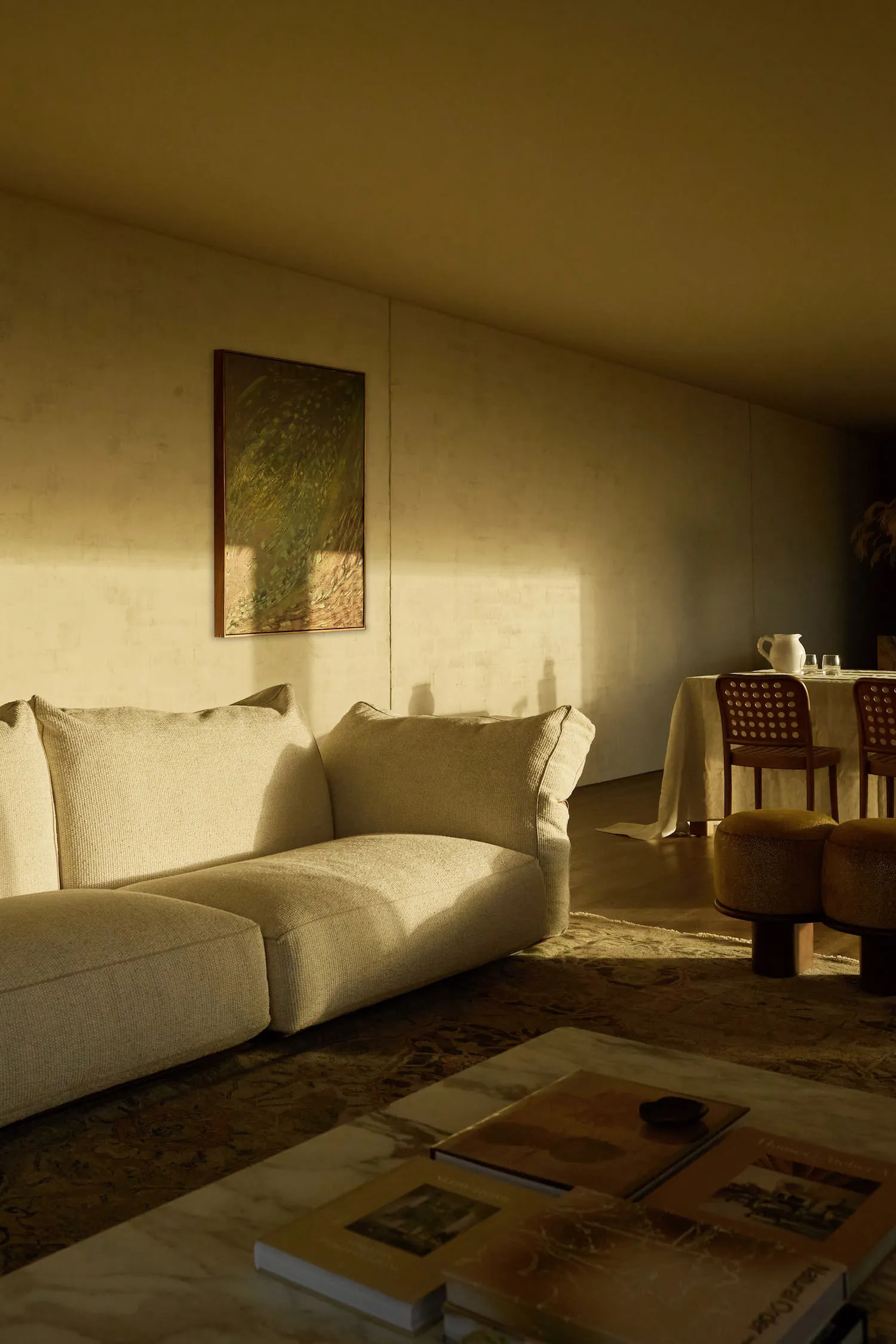 Minimalist living room featuring a cream bouclé sectional sofa, marble coffee table with art books, textured rug, and warm sunlight creating dramatic shadows on a textured wall with a landscape painting.