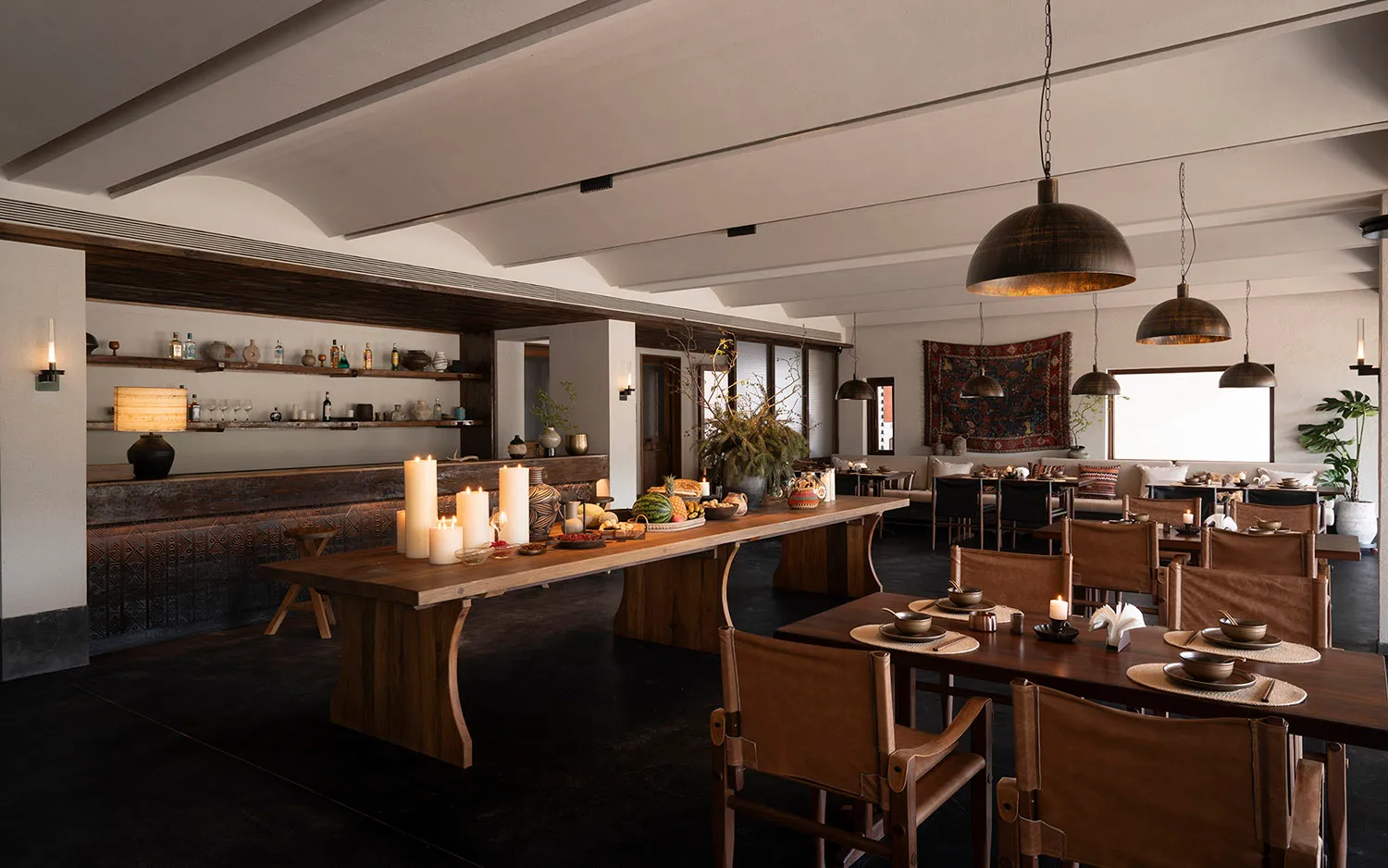 Spacious open-plan restaurant interior featuring a massive reclaimed wood communal table, brown leather sling chairs, textured dark walls, and layered tribal decor under vaulted ceilings.