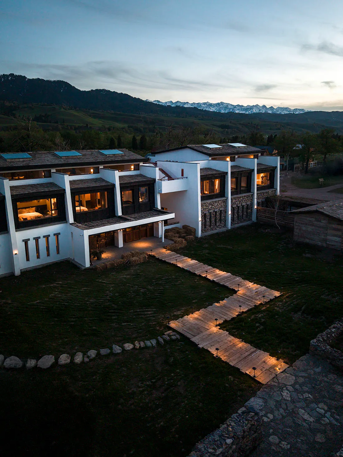 Aerial twilight view of a modern farmhouse-style resort villa featuring white walls, dark timber beams, and stone masonry, connected by illuminated wooden pathways set against rolling hills and snow-capped mountains.