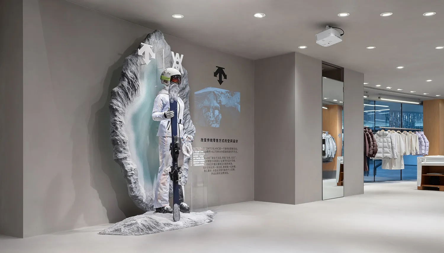 Modern retail space featuring a large textured artificial rock wall with a mannequin dressed in white ski gear holding skis, accompanied by branded graphics and clothing racks in the background.