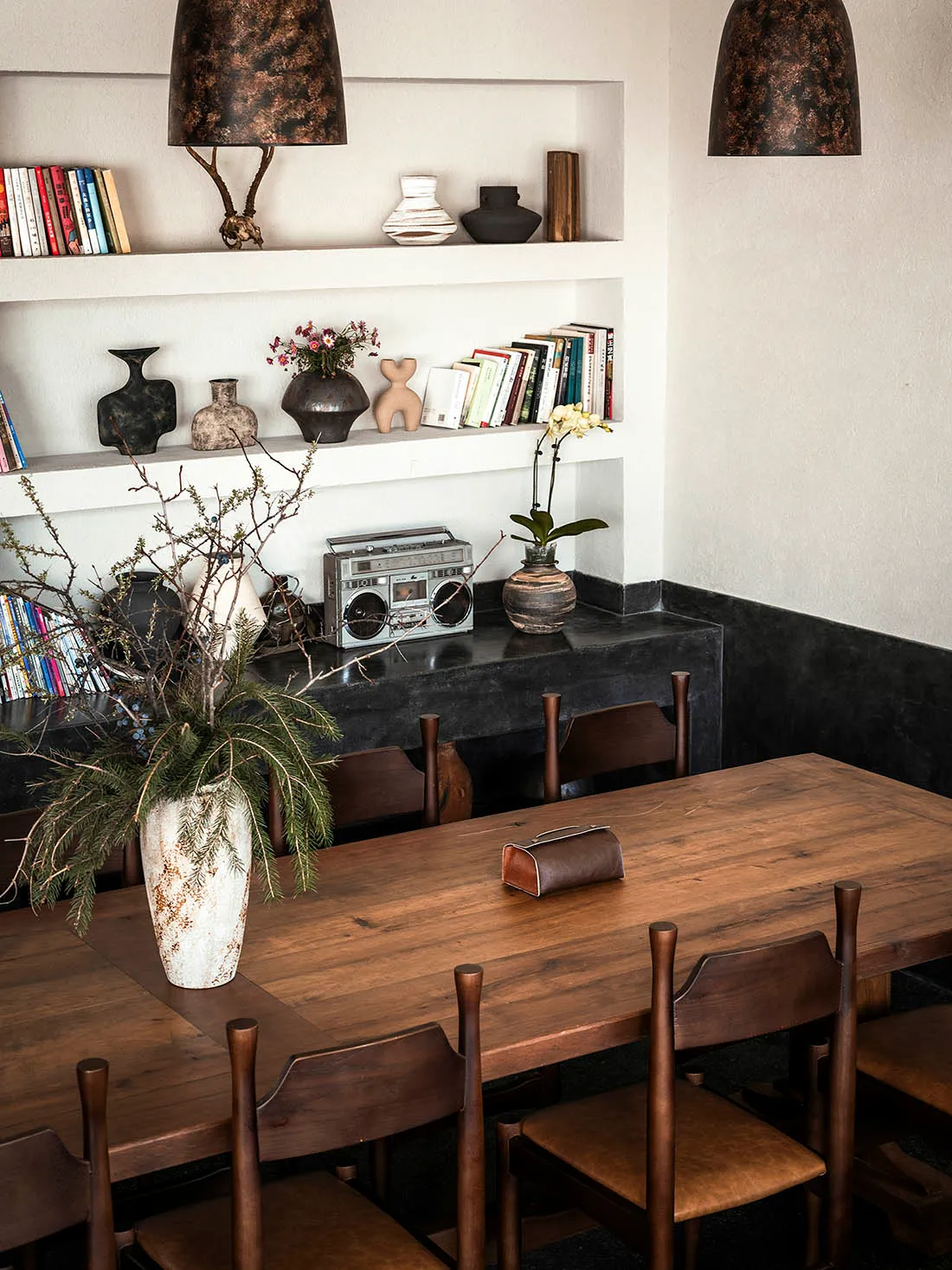 Warm dining room featuring a long reclaimed wood table, dark wooden spindle-back chairs, built-in white shelves with books and ceramics, and a dark plaster lower wall niche.