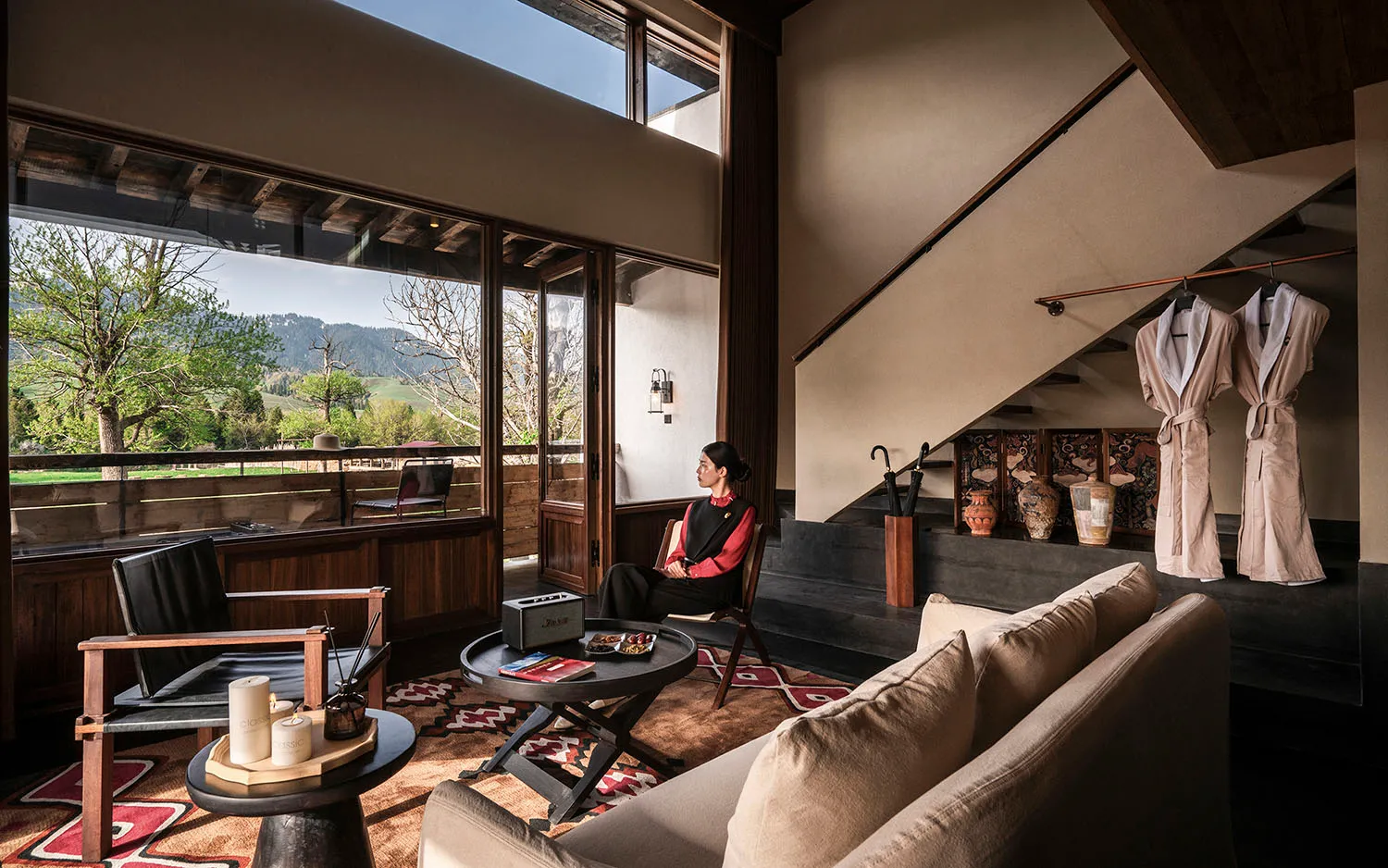 Modern hotel lounge with walnut wood paneling, beige sofa, black leather armchair, round coffee tables on a patterned rug, large glass windows overlooking green mountains, and a woman in traditional attire sitting by the staircase.