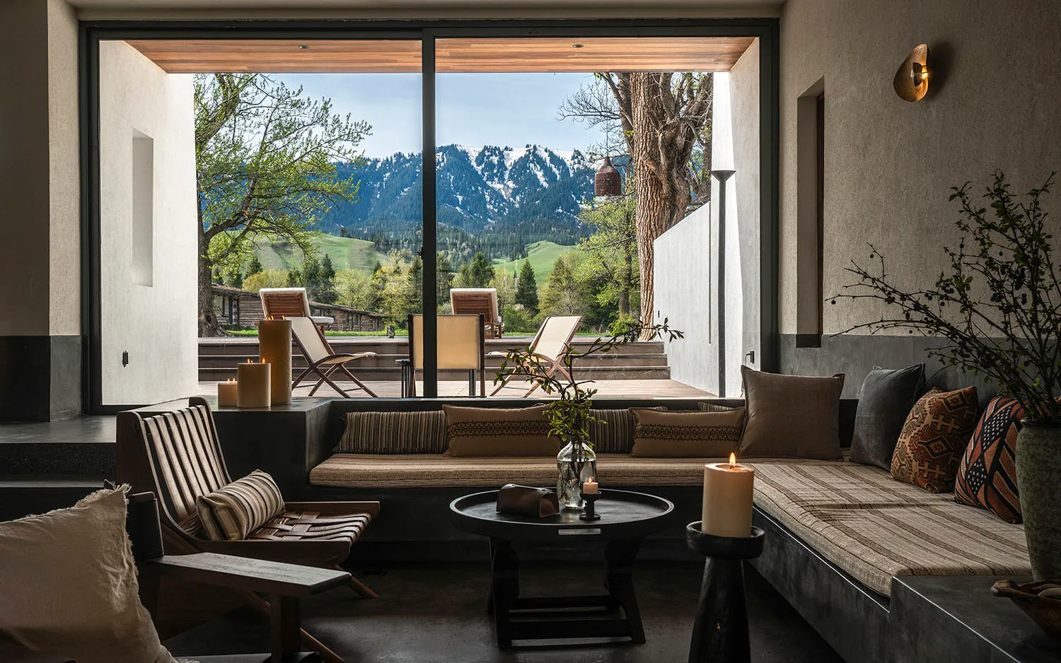 Modern minimalist living room with concrete walls, built-in striped banquette, wooden lounge chair, round coffee table, and large glass opening revealing snow-capped mountains and forest.