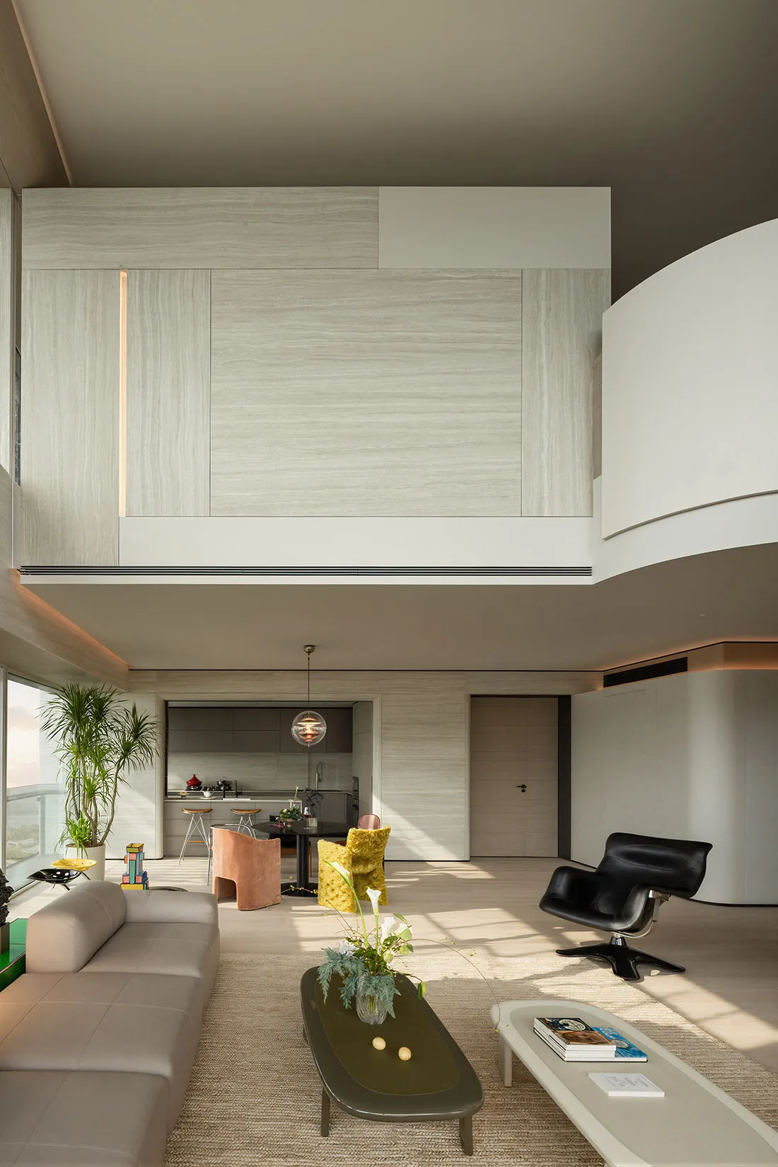 Modern double-height living room featuring light wood paneling, a curved white wall, beige sectional sofa, black leather lounge chair, and colorful yellow and terracotta accent chairs in an open-plan layout.