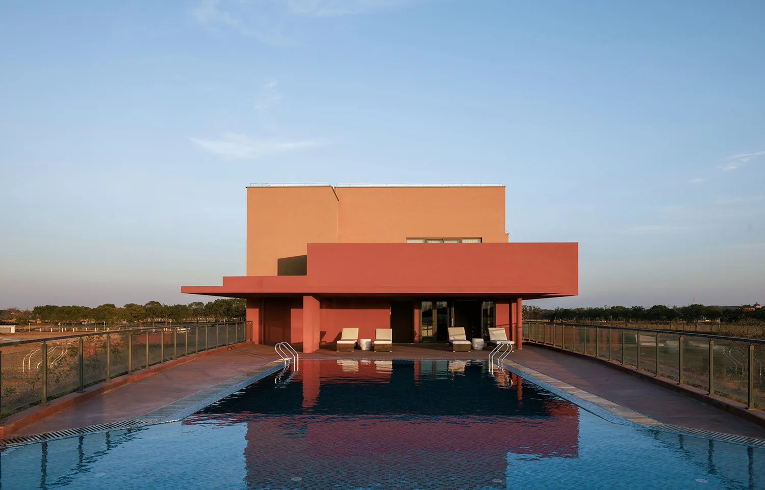 Modern terracotta-colored villa with a long rectangular rooftop swimming pool, four lounge chairs, and glass railings overlooking a rural landscape at dusk.
