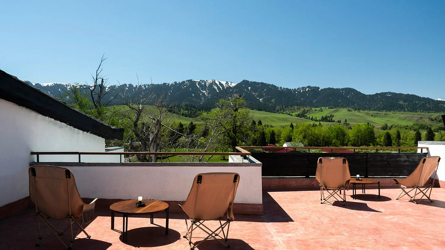 Outdoor rooftop terrace with four tan canvas folding chairs, round wooden side tables, terracotta tiled floor, and panoramic mountain landscape under a clear blue sky.