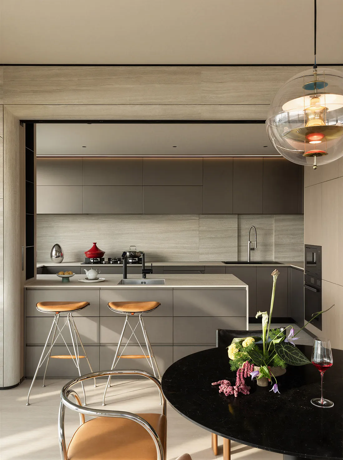 Modern minimalist kitchen with grey matte cabinetry, central island with two leather bar stools, and a round black marble dining table set with a vibrant floral arrangement and red wine.