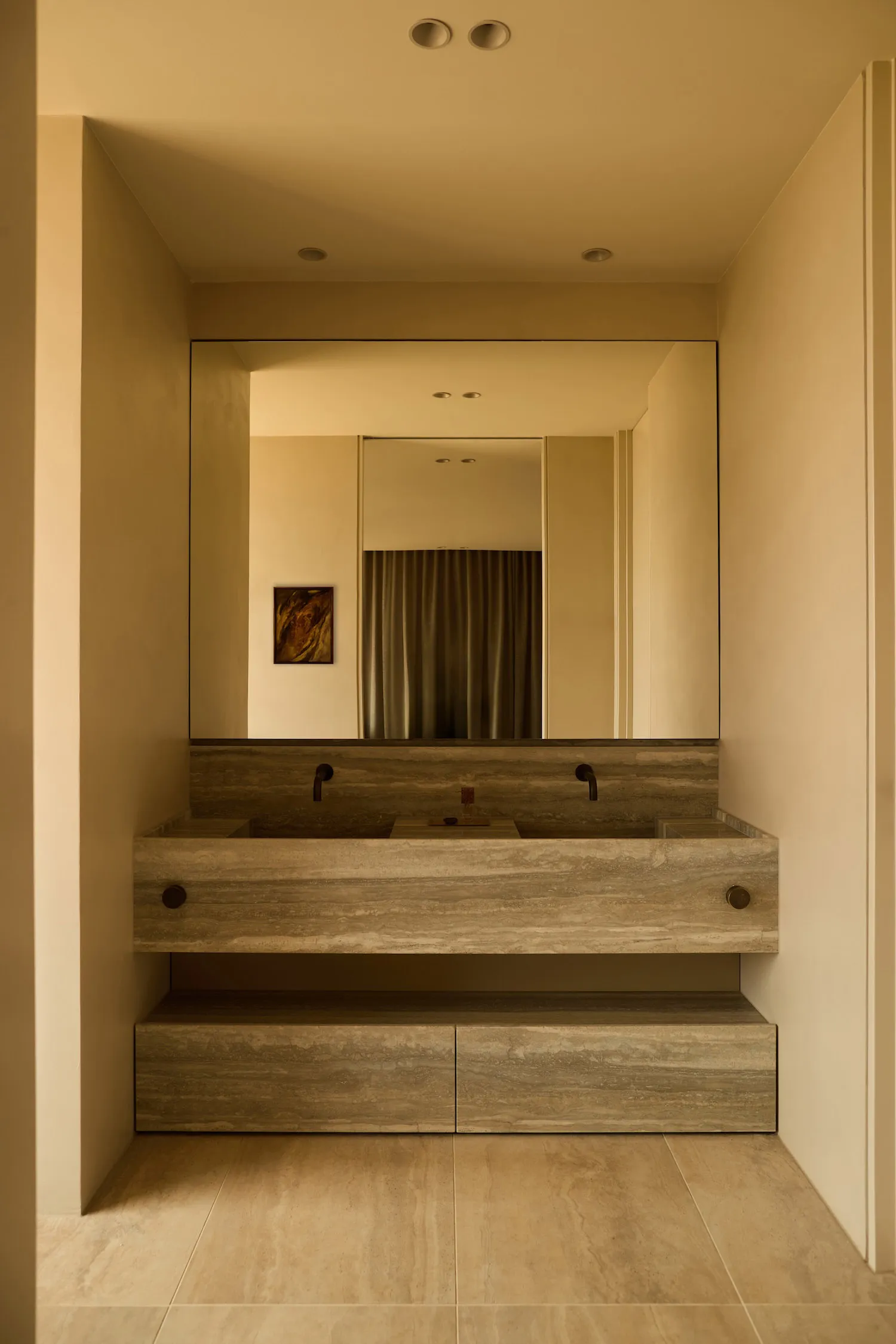 Modern minimalist bathroom with a long floating travertine double vanity, two black wall-mounted faucets, large frameless mirror, and warm beige tiled floor