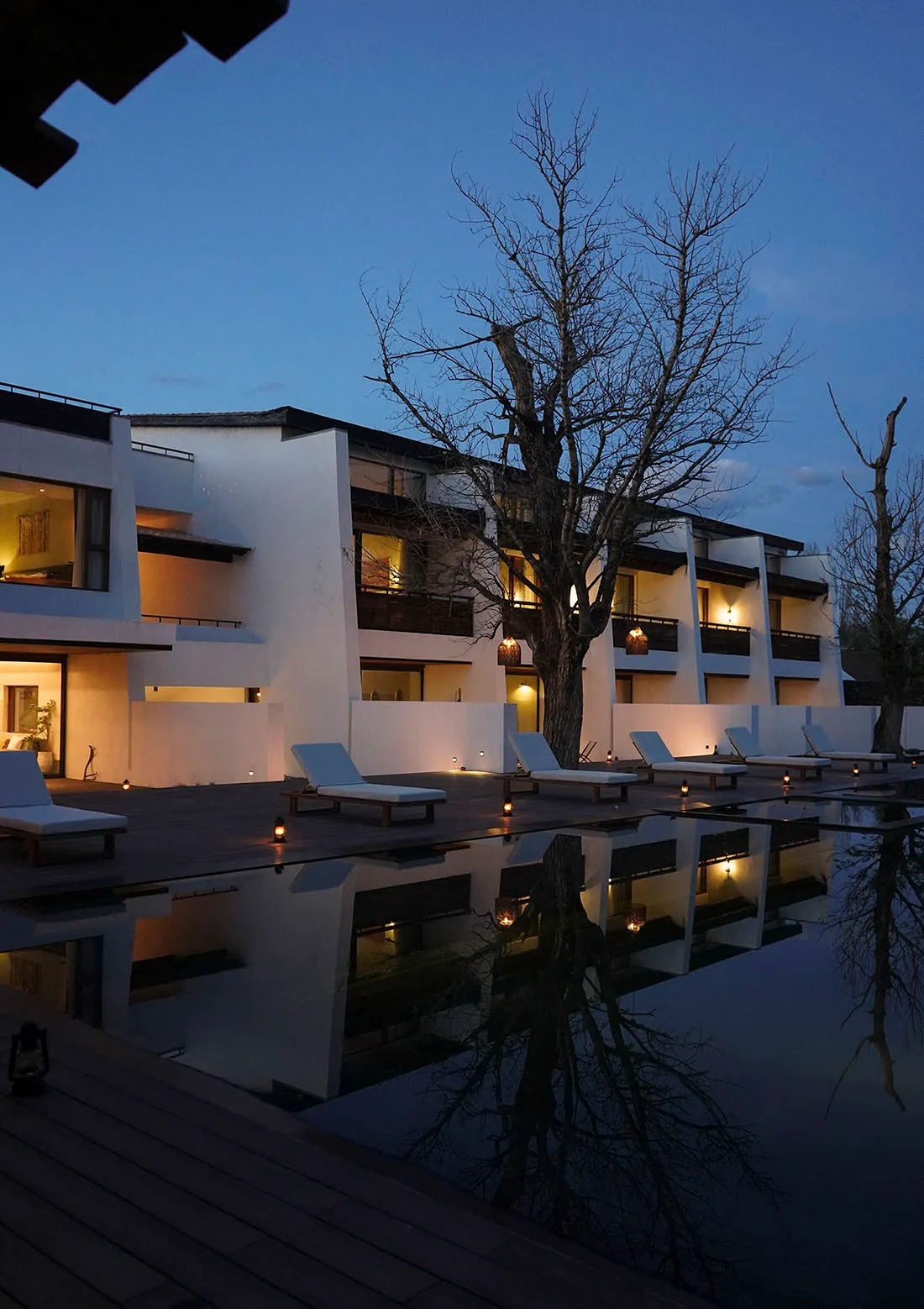Modern resort pool deck at twilight with white lounge chairs, wooden deck, reflecting pool, and illuminated minimalist white buildings with bare trees against a deep blue sky.