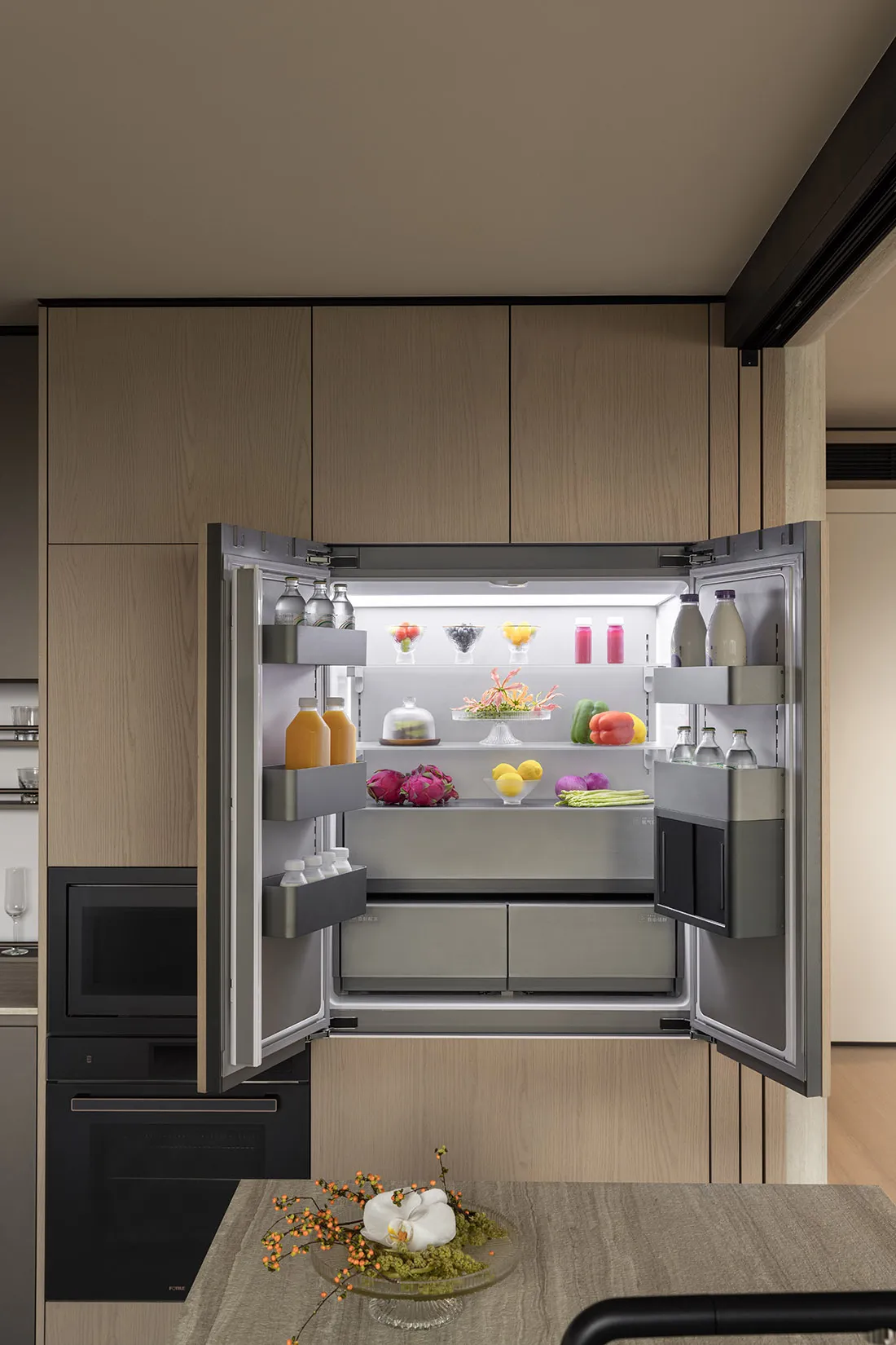 Modern kitchen featuring a large built-in refrigerator with both doors open, displaying fresh fruits, vegetables, and beverages on illuminated shelves, surrounded by light oak cabinetry.