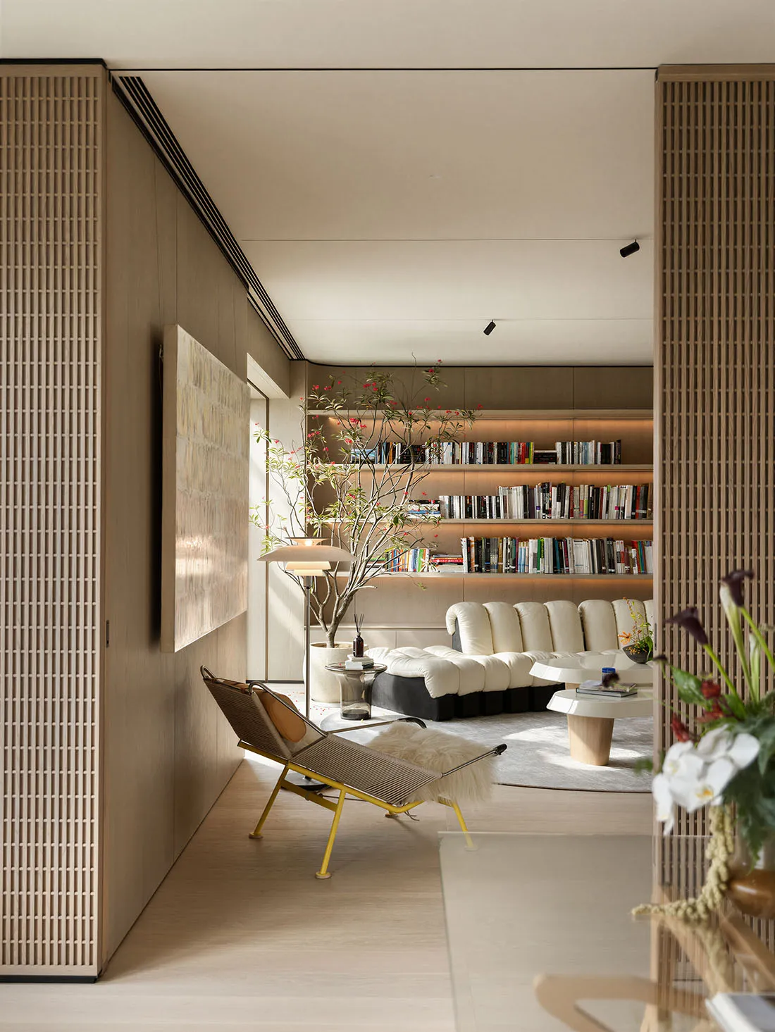 Modern minimalist living room featuring built-in walnut bookshelves, a white modular sofa, a yellow metal and rattan lounge chair with sheepskin throw, and a tall potted tree under soft natural lighting.