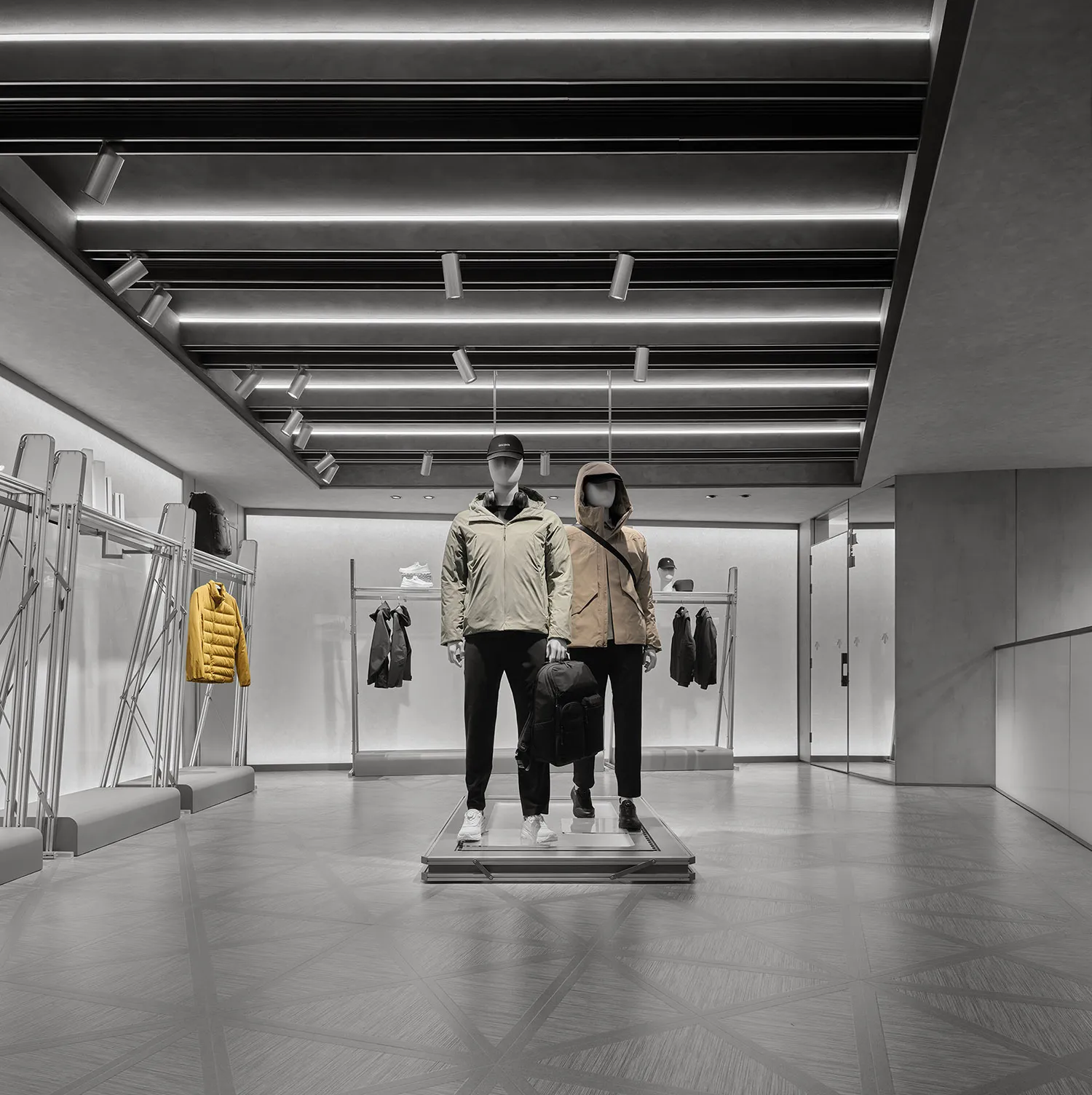 Modern minimalist menswear retail store interior with raw concrete walls, geometric floor tiles, linear LED ceiling lights, and two mannequins dressed in contemporary jackets displayed on a central metal platform.