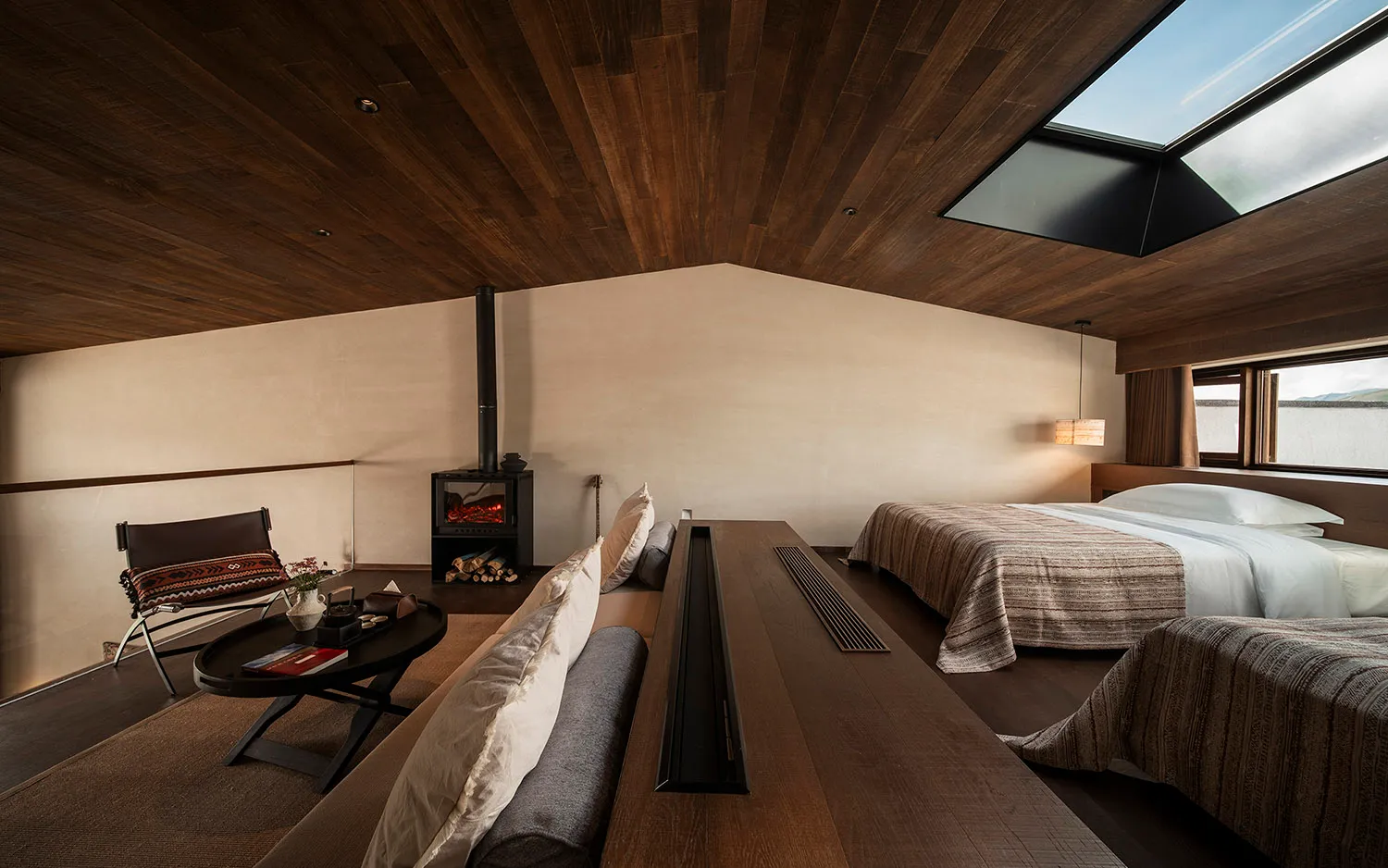 Modern loft-style bedroom with dark wood ceiling, large skylights, wood-burning stove, and two beds with neutral striped bedding.