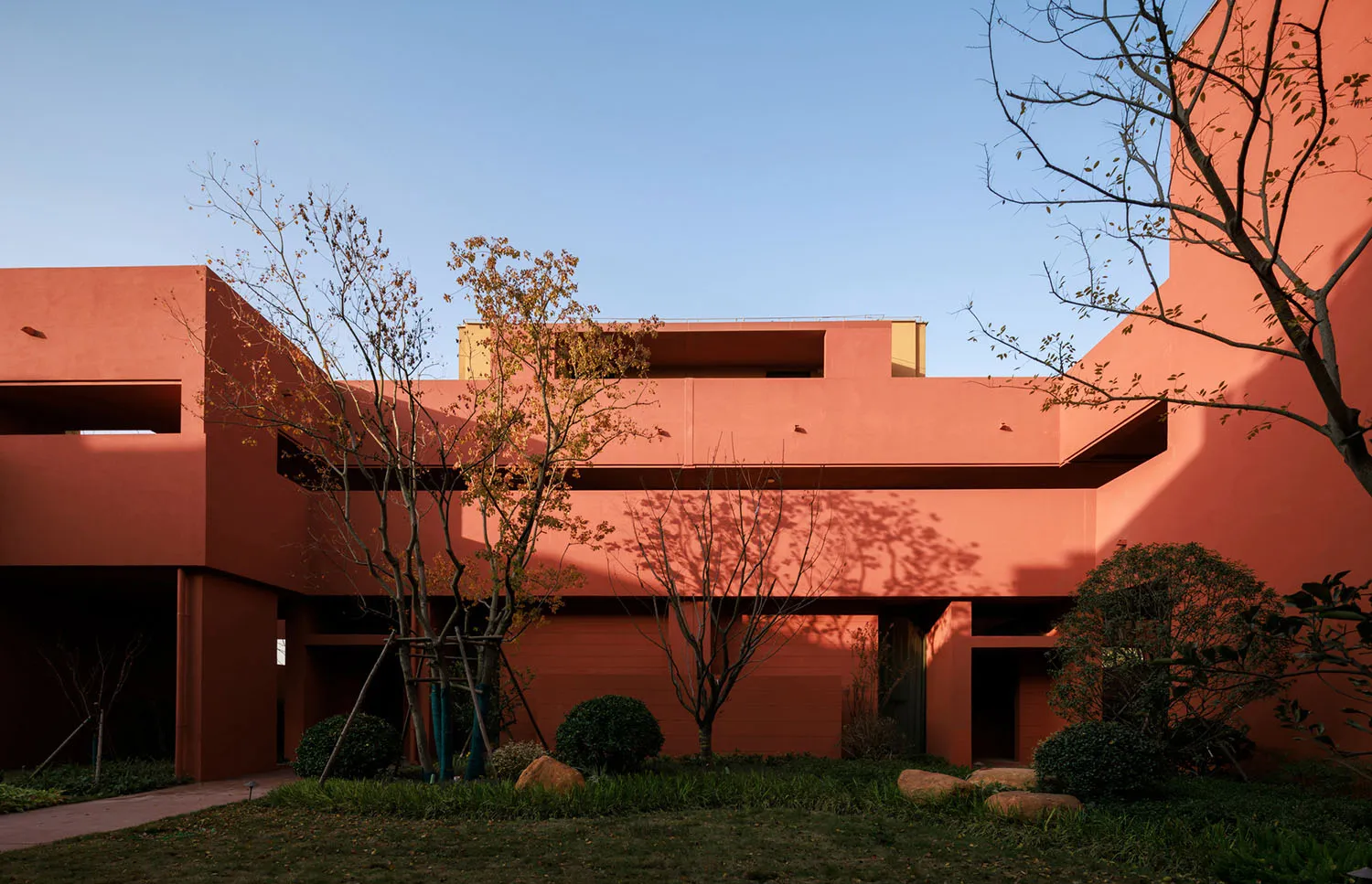 Exterior view of a modern terracotta-colored building with geometric forms, surrounded by a landscaped courtyard with trees and shrubs under a clear blue sky.
