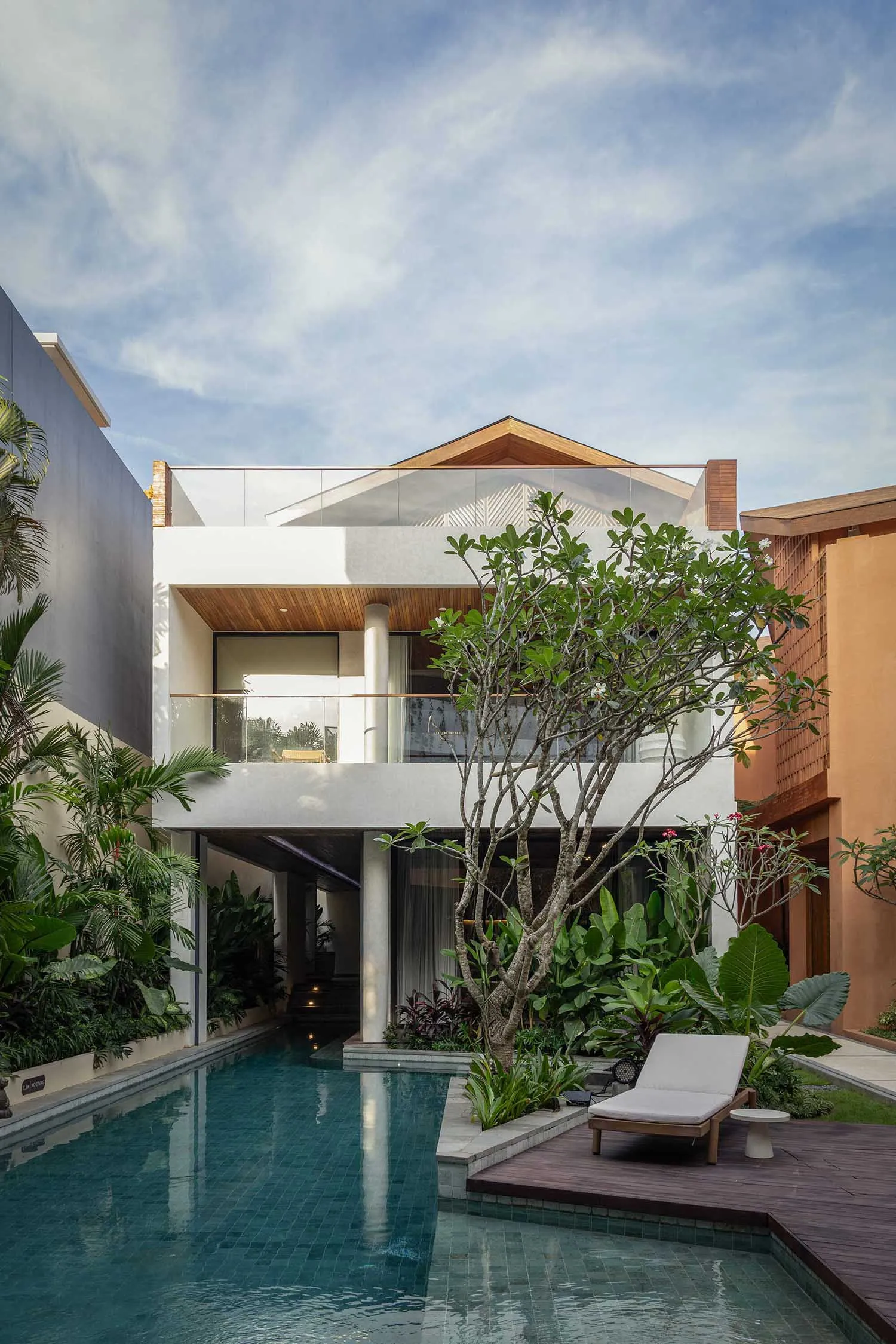 Modern tropical villa with a long lap pool in a lush courtyard, featuring concrete walls, warm wood ceilings, and abundant tropical plants under a bright blue sky.