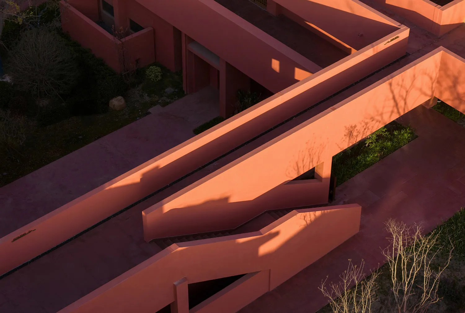 Aerial view of a modern terracotta-colored architectural staircase and concrete walkways bathed in warm golden sunset light, casting strong geometric shadows.