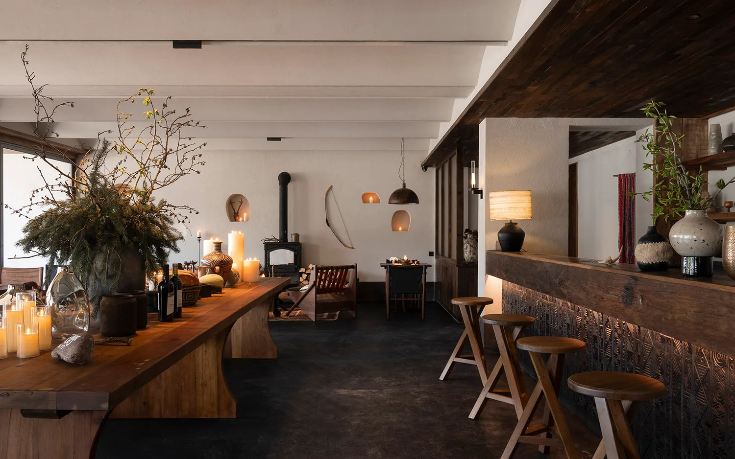 Spacious open-plan rustic modern living and dining room featuring a long reclaimed wood dining table, wooden bar counter with three stools, black wood-burning stove, and abundant natural textures and candlelight.