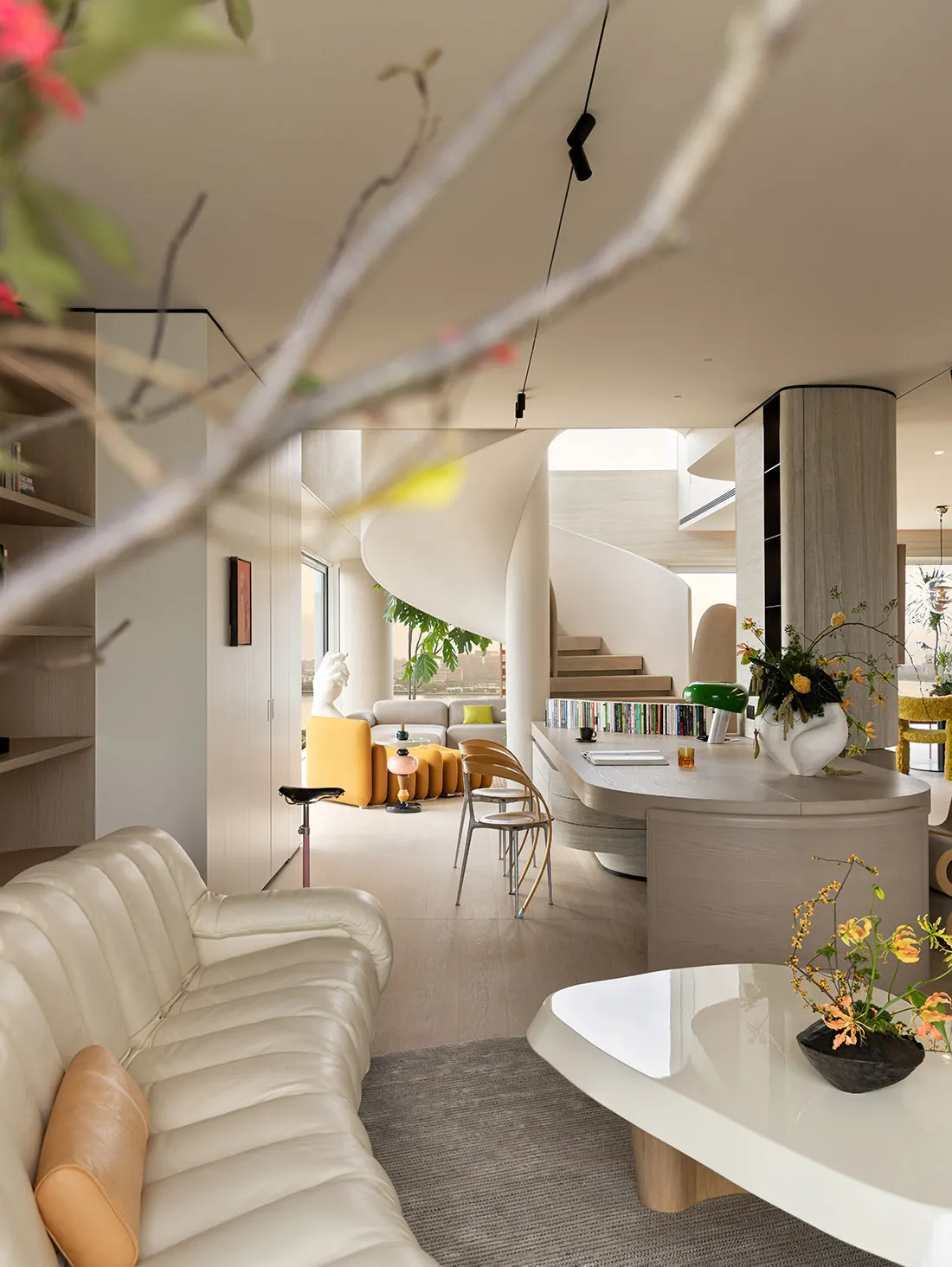 Modern open-concept living room featuring a cream ribbed leather sofa, curved wooden kitchen island, dramatic white spiral staircase, and vibrant yellow furniture in the background.