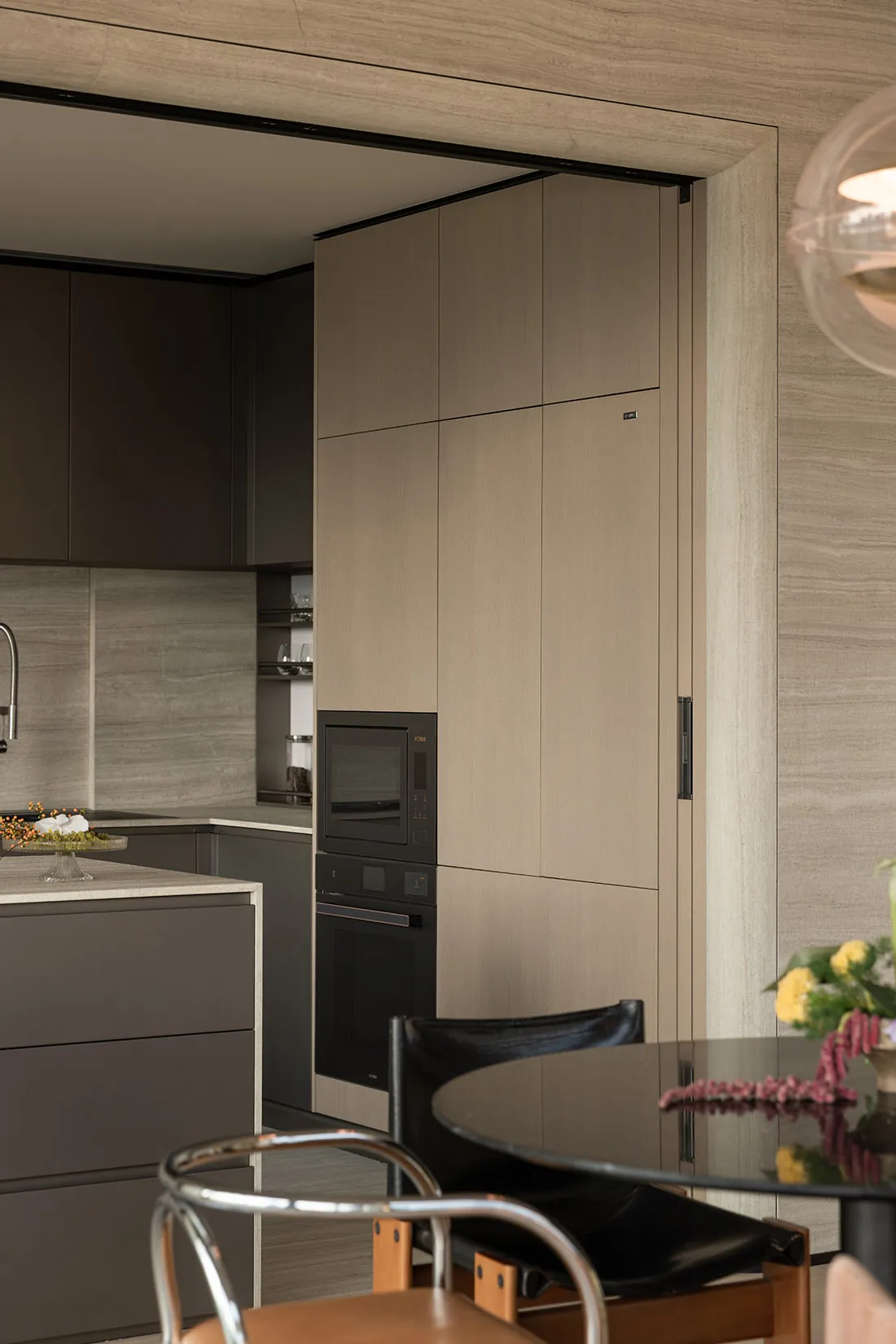 Modern minimalist kitchen featuring taupe wood-grain handleless cabinets, integrated black oven and microwave, marble-look backsplash, and a black leather dining chair in the foreground.