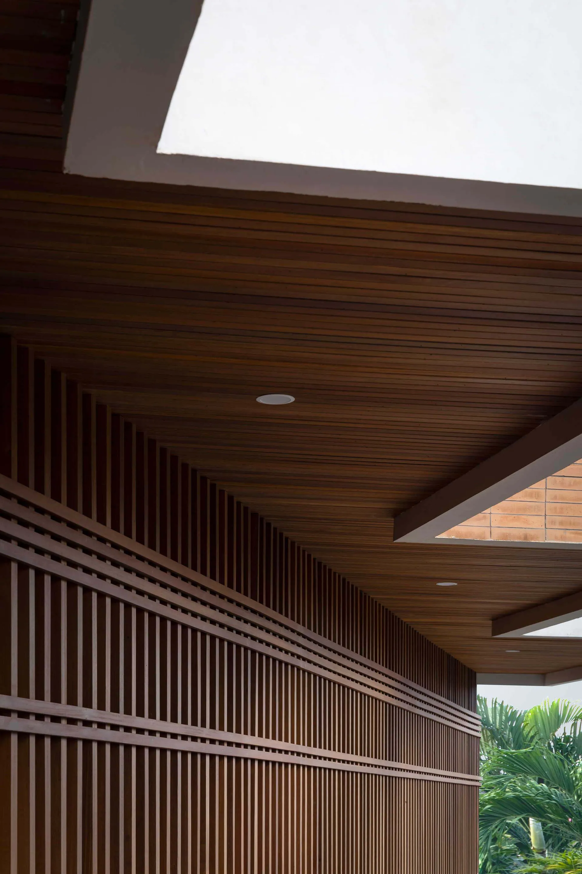 Low-angle view of a modern outdoor corridor featuring a dark teak wood slatted ceiling with recessed lighting and tropical plants visible on the side.