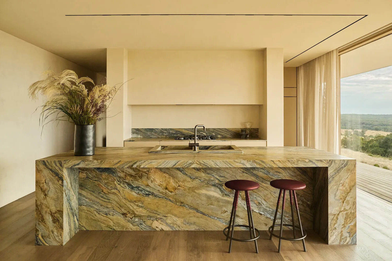 Modern kitchen featuring a massive onyx marble island with dramatic veining, integrated sink, two burgundy leather bar stools, and large windows overlooking a natural landscape.