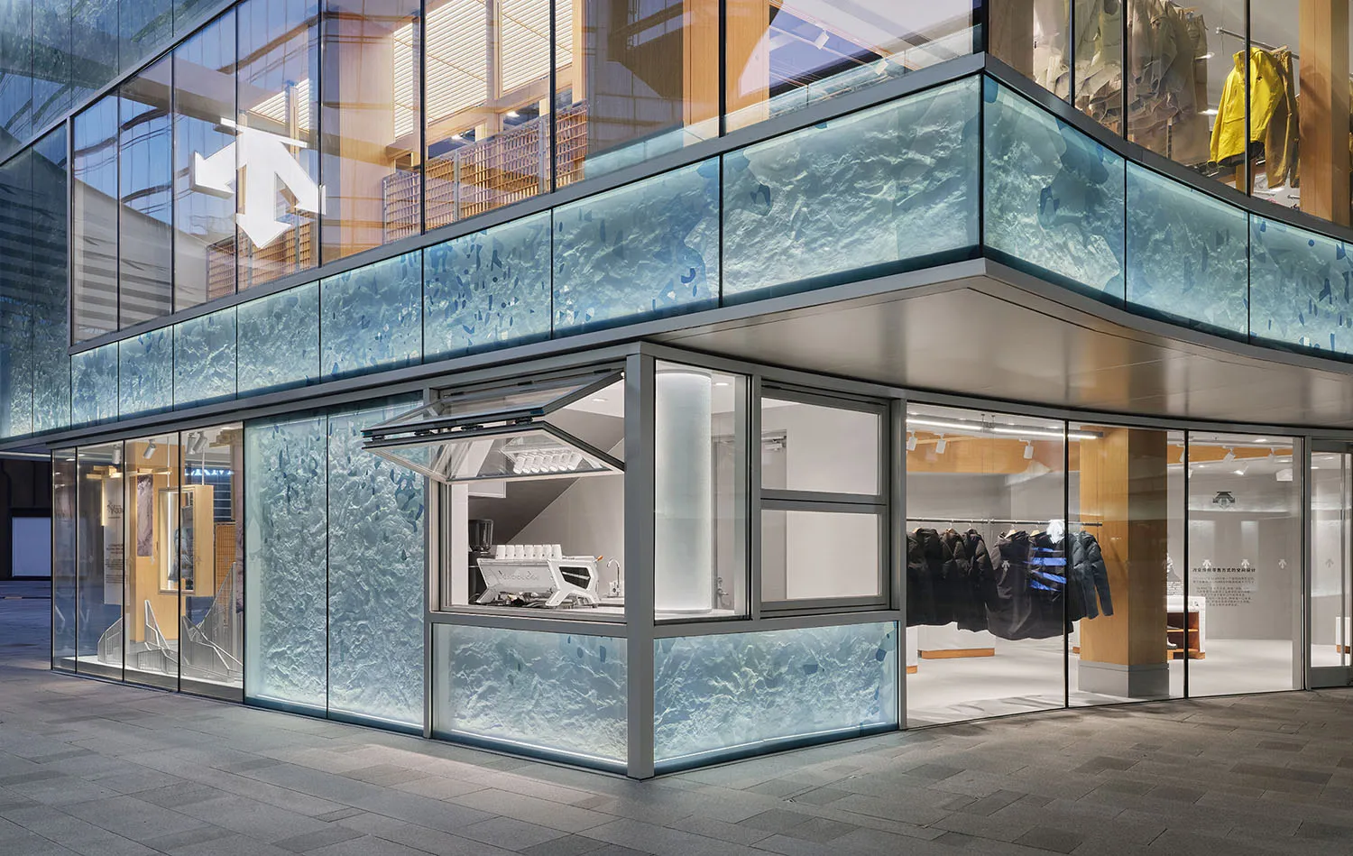 Modern retail store exterior at night with large blue textured cast-glass panels resembling ice, large glass windows, and visible clothing displays inside.