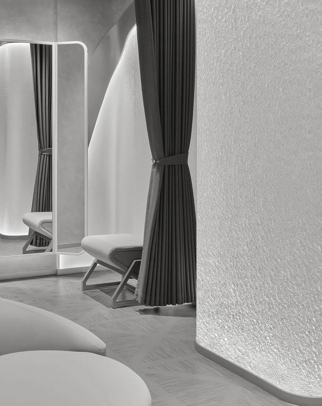 Modern minimalist dressing room featuring curved textured plaster walls, dark grey pleated curtains, white leather bench, and a tall backlit mirror with LED lighting.