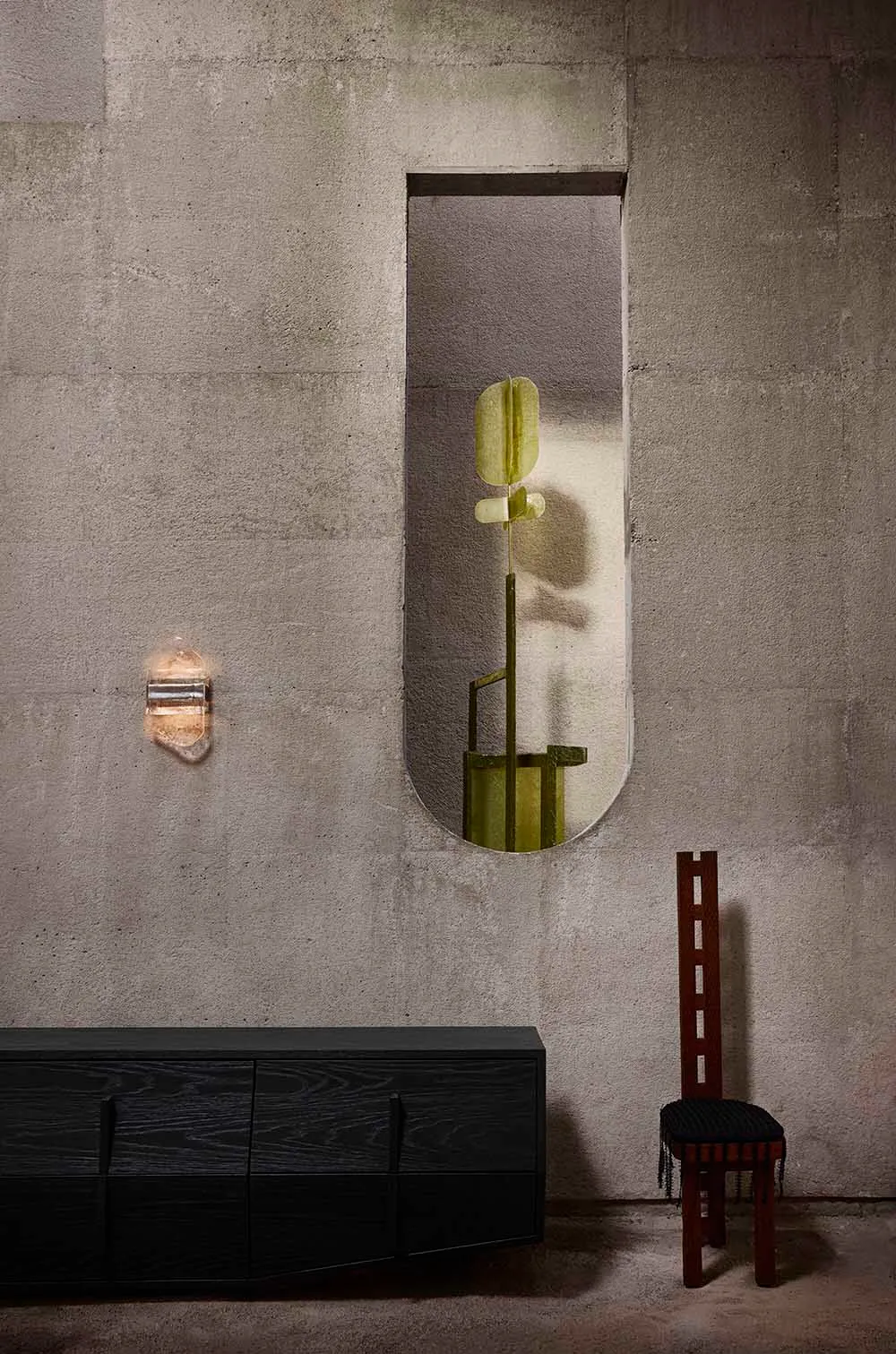 Minimalist brutalist interior featuring raw concrete walls, a black charred wood console, a tall dark wood chair with black cushion, a modern wall sconce, and an olive-green abstract sculpture viewed through a vertical oval opening.