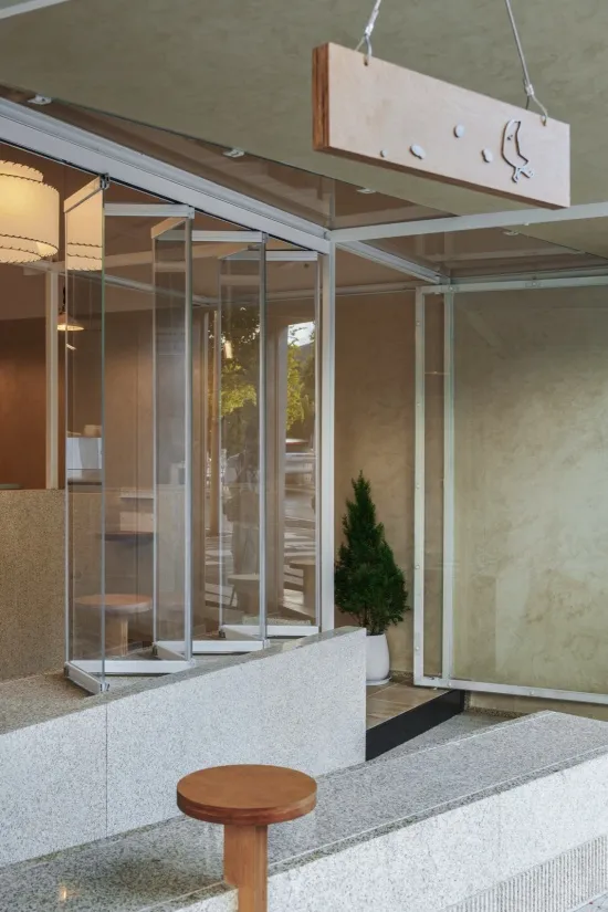 Modern cafe entrance featuring speckled terrazzo counters, wooden stools, a folding glass partition system, and a hanging wooden sign with bird logo.