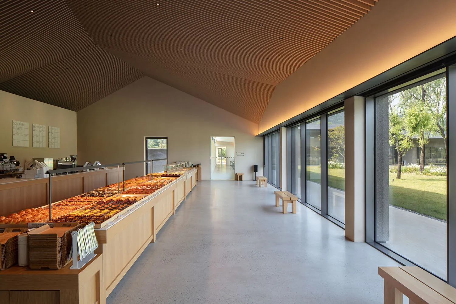 Modern minimalist bakery interior with a long oak pastry display counter, polished concrete floor, slatted wooden ceiling, and large glass windows facing a green garden.