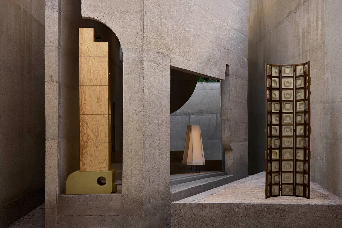 Modern brutalist interior courtyard featuring raw concrete walls, a tall oak wood screen, a folding patterned room divider, and a lit conical floor lamp on stepped platforms.