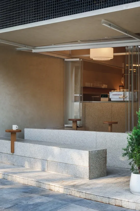 Modern minimalist cafe exterior featuring a long terrazzo bench, wooden stools, concrete walls, and a pendant lamp visible inside.