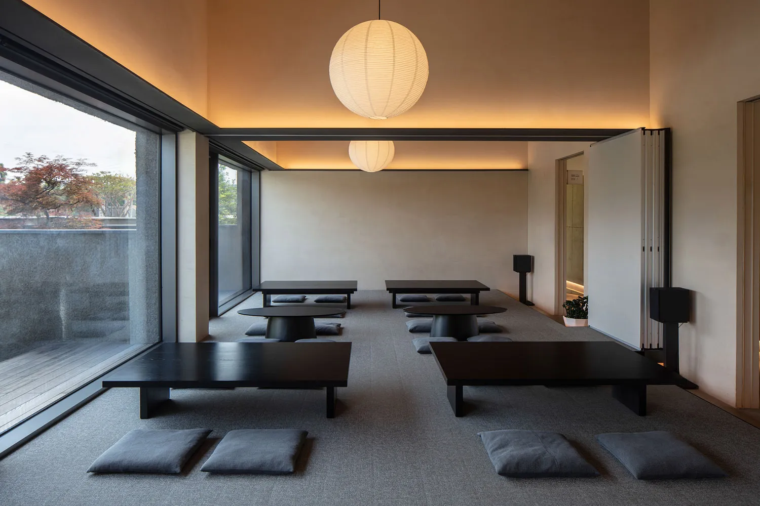 Modern Japanese-style meditation room with dark low tables, grey floor cushions, large paper lanterns, and floor-to-ceiling windows overlooking greenery.