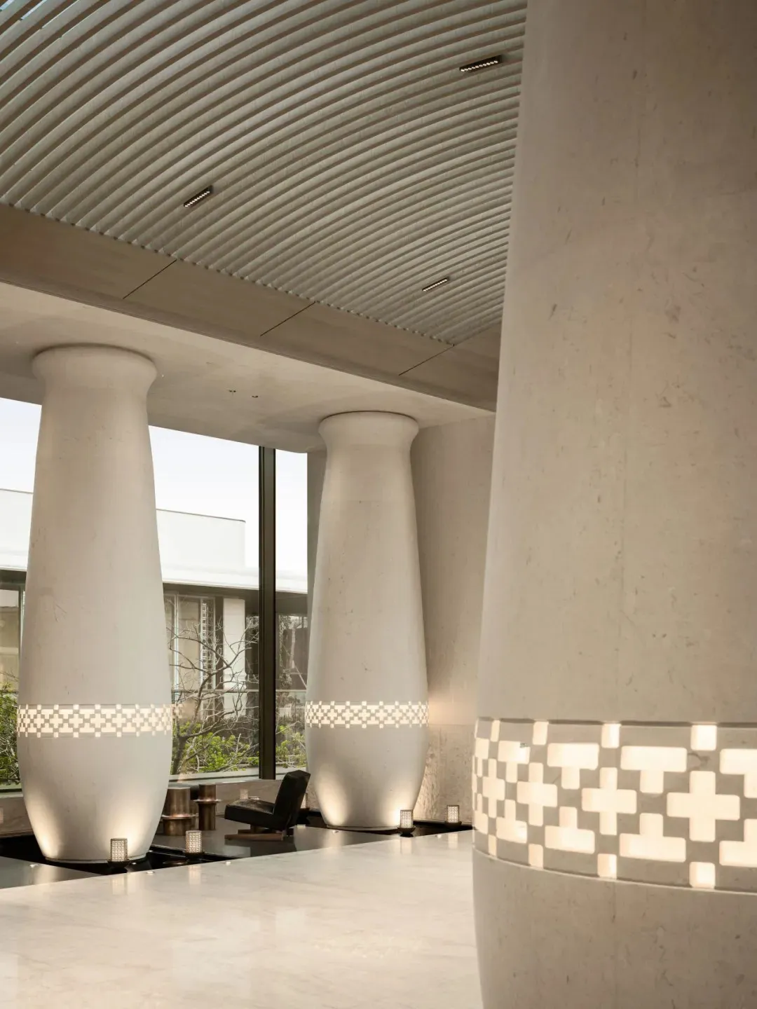Modern minimalist lobby featuring three large tapered travertine columns with illuminated geometric tile bands, ribbed concrete ceiling, and polished stone floor.