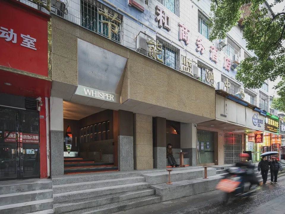 Street view of modern brutalist restaurant entrance with wide stone steps, recessed lighting, and a large travertine panel bearing the word WHISPER.