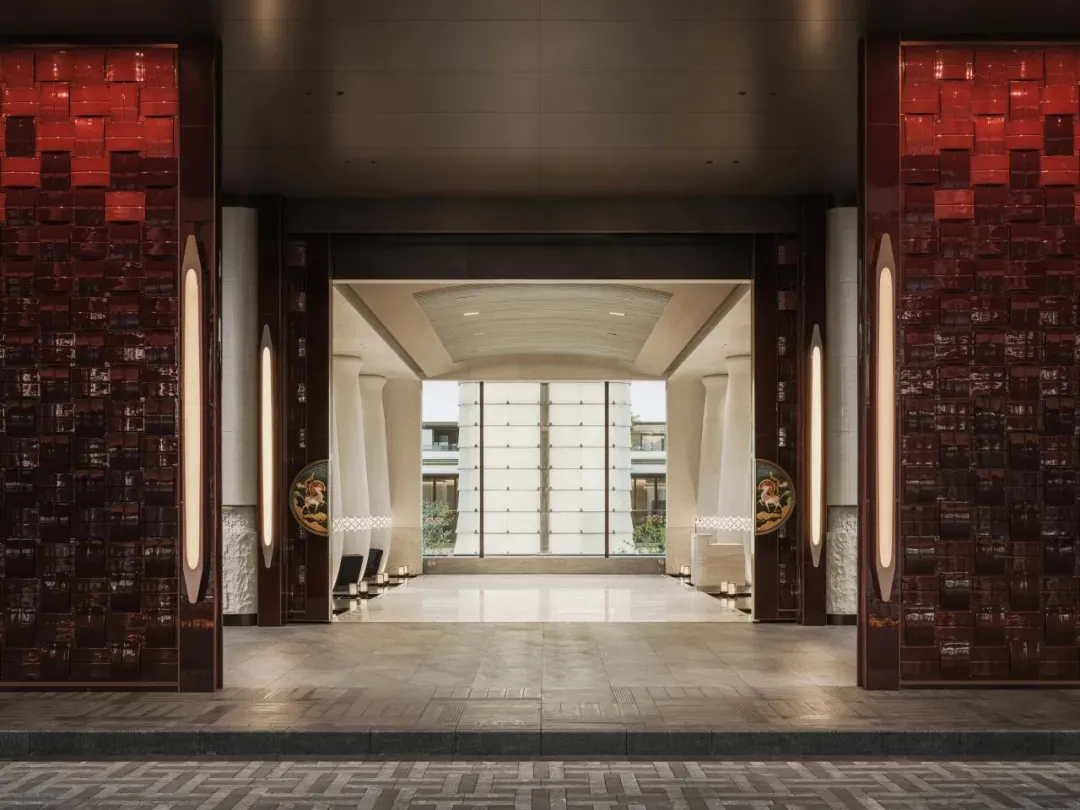 Symmetrical view of a luxurious hotel lobby entrance featuring deep red textured tiled walls, vertical light fixtures, circular art panels with horse motifs, white columns, and a bright courtyard visible in the background.