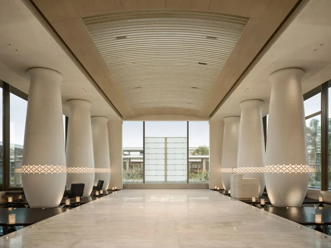Long symmetrical hotel lobby hallway featuring large white travertine columns with glowing patterned light bands, polished marble floor, and dramatic curved ceiling.