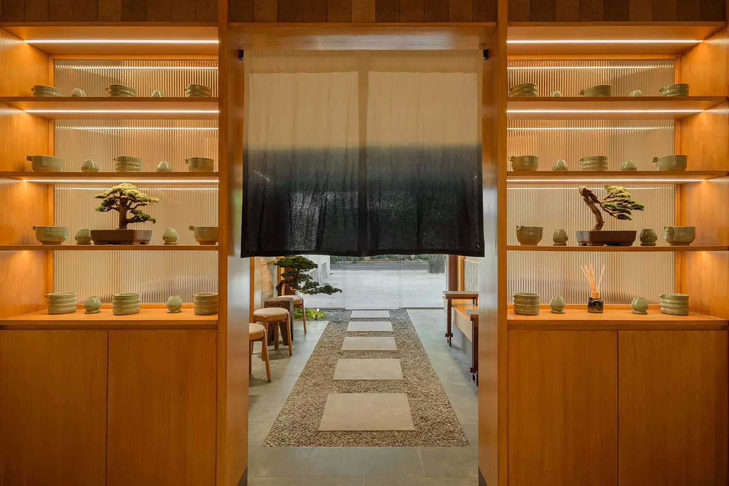 View through a Japanese tea room entrance with backlit oak display shelves filled with ceramic bowls and bonsai trees, leading to a gravel garden path with noren curtain.