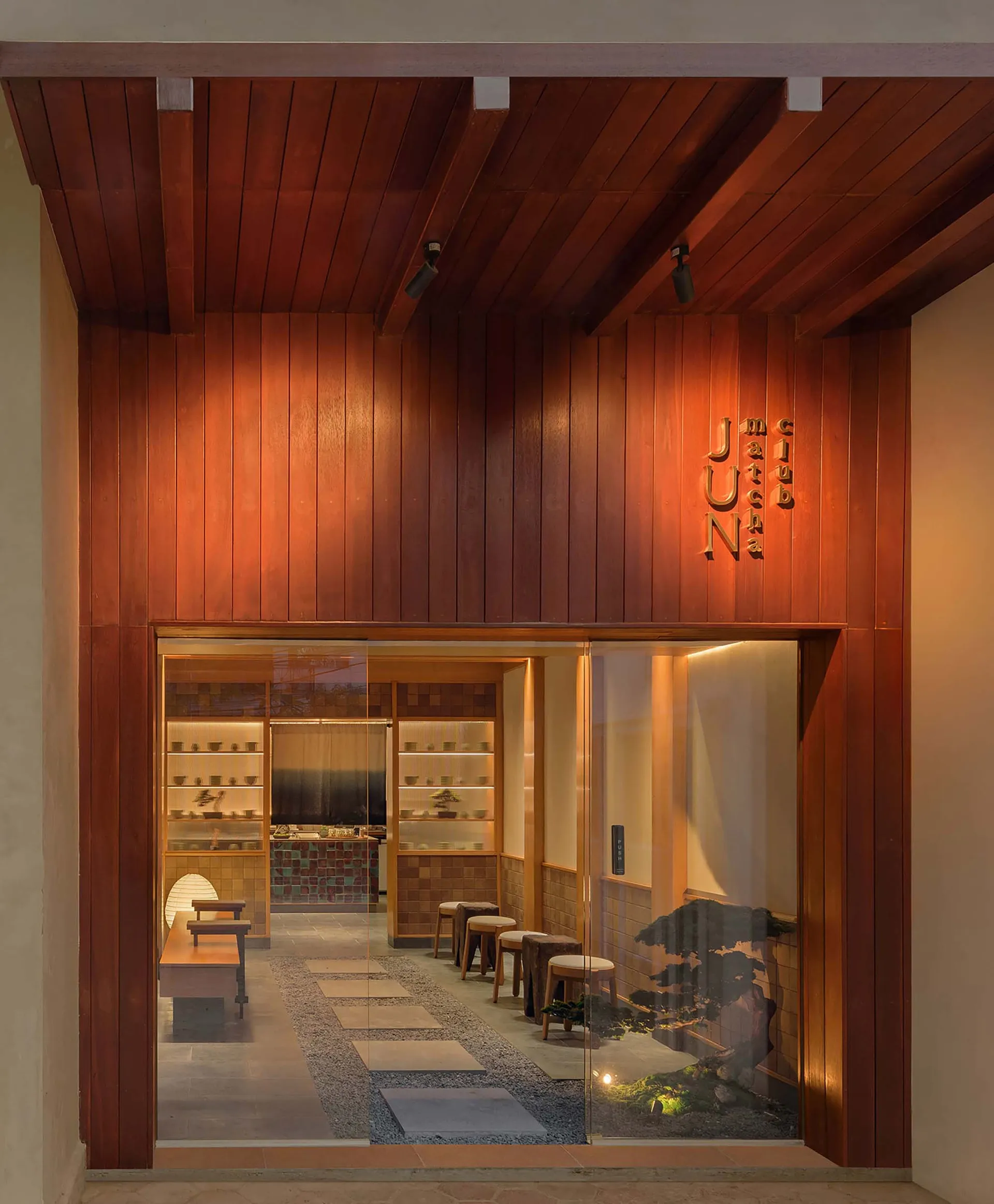 Modern Japanese tea room interior with rich reddish-brown wood paneling, stone pathway, low wooden stools, and a glass entrance featuring a bonsai garden.