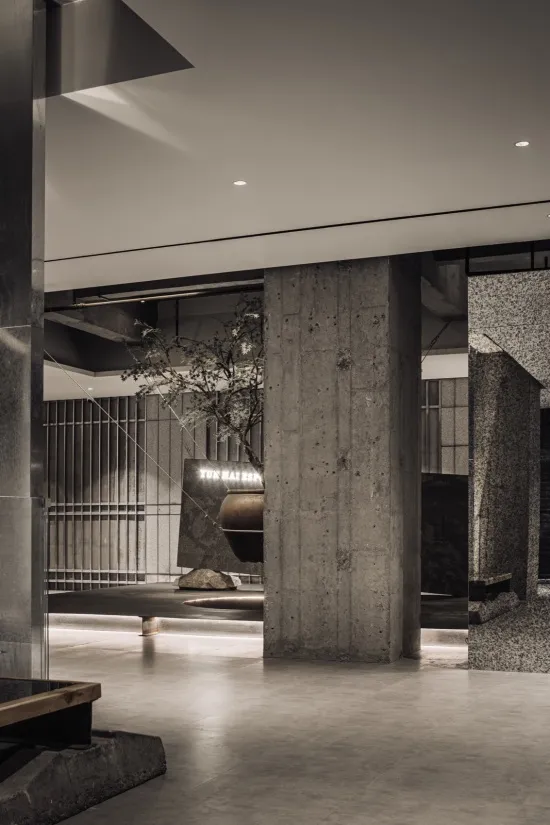 Modern brutalist interior lobby featuring massive raw concrete pillars, terrazzo accents, a large stone bonsai tree, and dramatic architectural lighting.