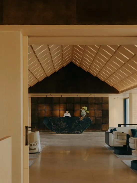 Modern luxury hotel lobby viewed from an entrance, featuring a black marble reception desk, warm wood-paneled ceiling with geometric folds, and contemporary seating area.