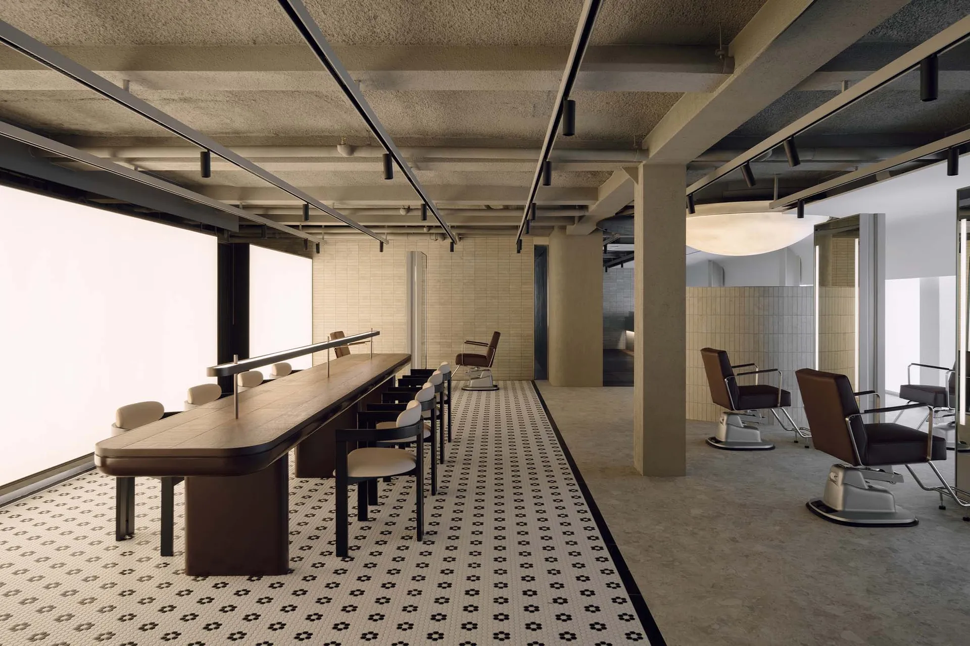 Modern brutalist hair salon interior featuring a long walnut styling counter, cream upholstered chairs, brown leather salon chairs on chrome bases, black and white patterned terrazzo floor, and raw concrete ceiling with linear lighting.