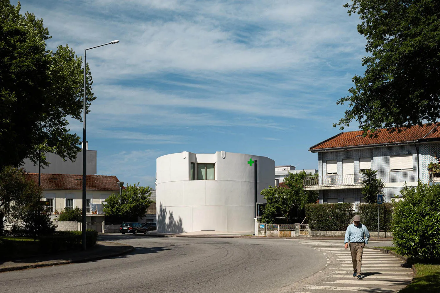 A modern white cylindrical building with minimal windows located at a street corner in a European residential neighborhood, with a man crossing the street in the foreground.