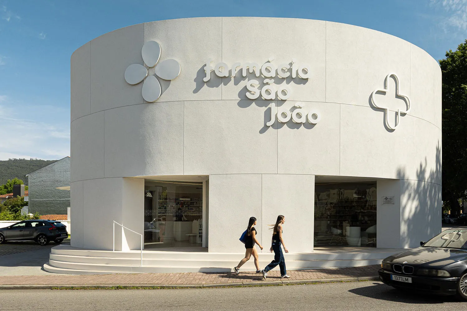 Modern cylindrical white pharmacy building with curved facade, large flower and cross signage, and glass storefront under bright daylight.