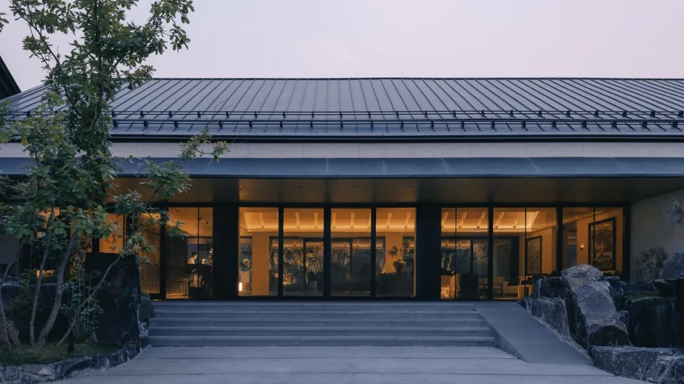 Modern single-story residence with floor-to-ceiling glass facade, concrete steps, and large natural stone landscaping at twilight.